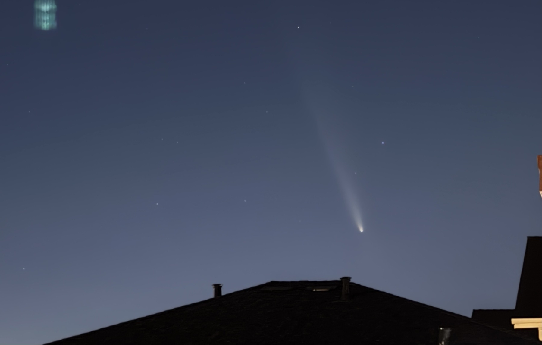 A bright point of light with a long hazy tail is low above a nearby roof.