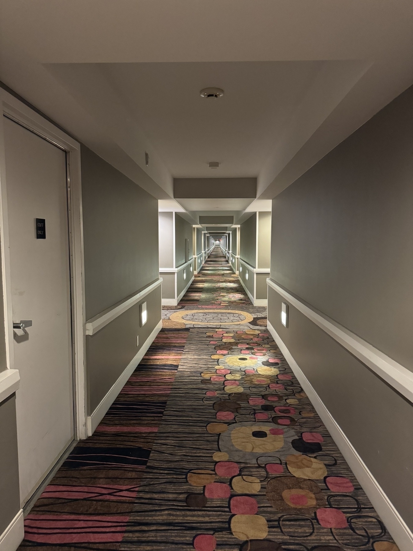 A very long, empty hotel hallway with 70s patterned carpet and grey walls. It looks like a render.