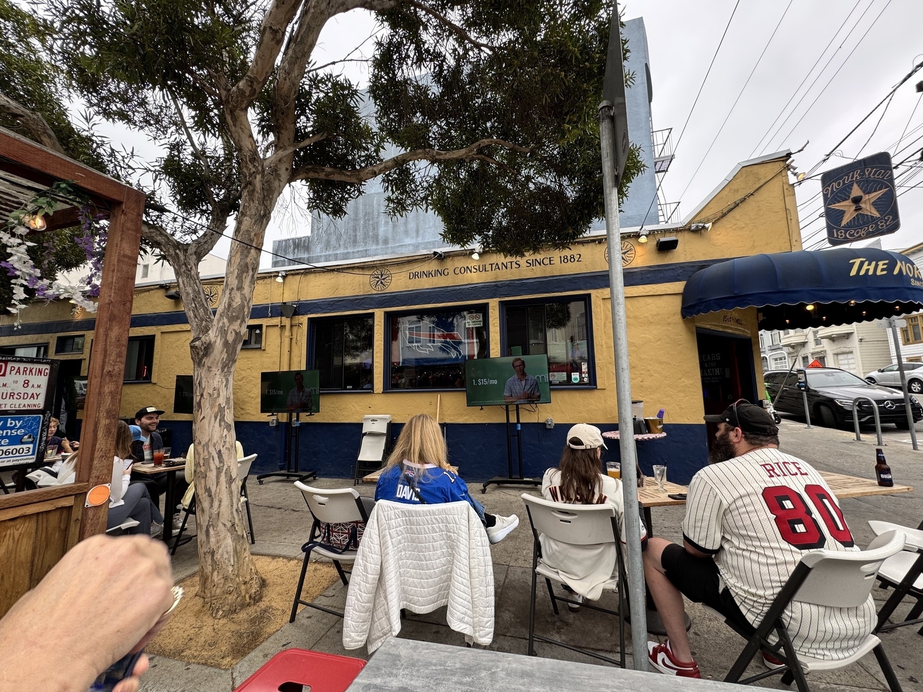 People sitting outside in front of large screen TVs showing the Buffalo Bills game. The bar is a small, local place called Northstar, and the sign above it says “drinking consultants since 1882”.