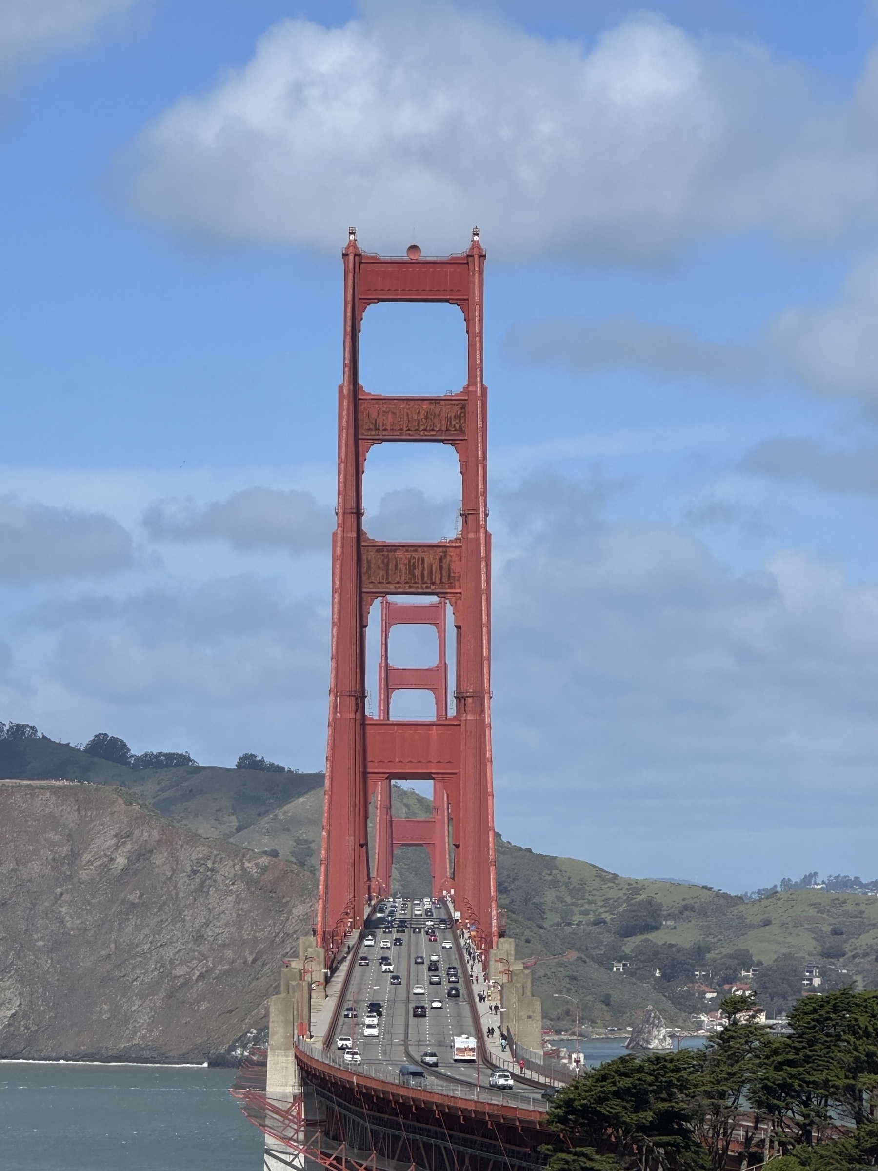 Looking down along the length of the Golden Gate Bridge against a blue-green bay, distant cliffs, and a partly cloudy, sunny sky.