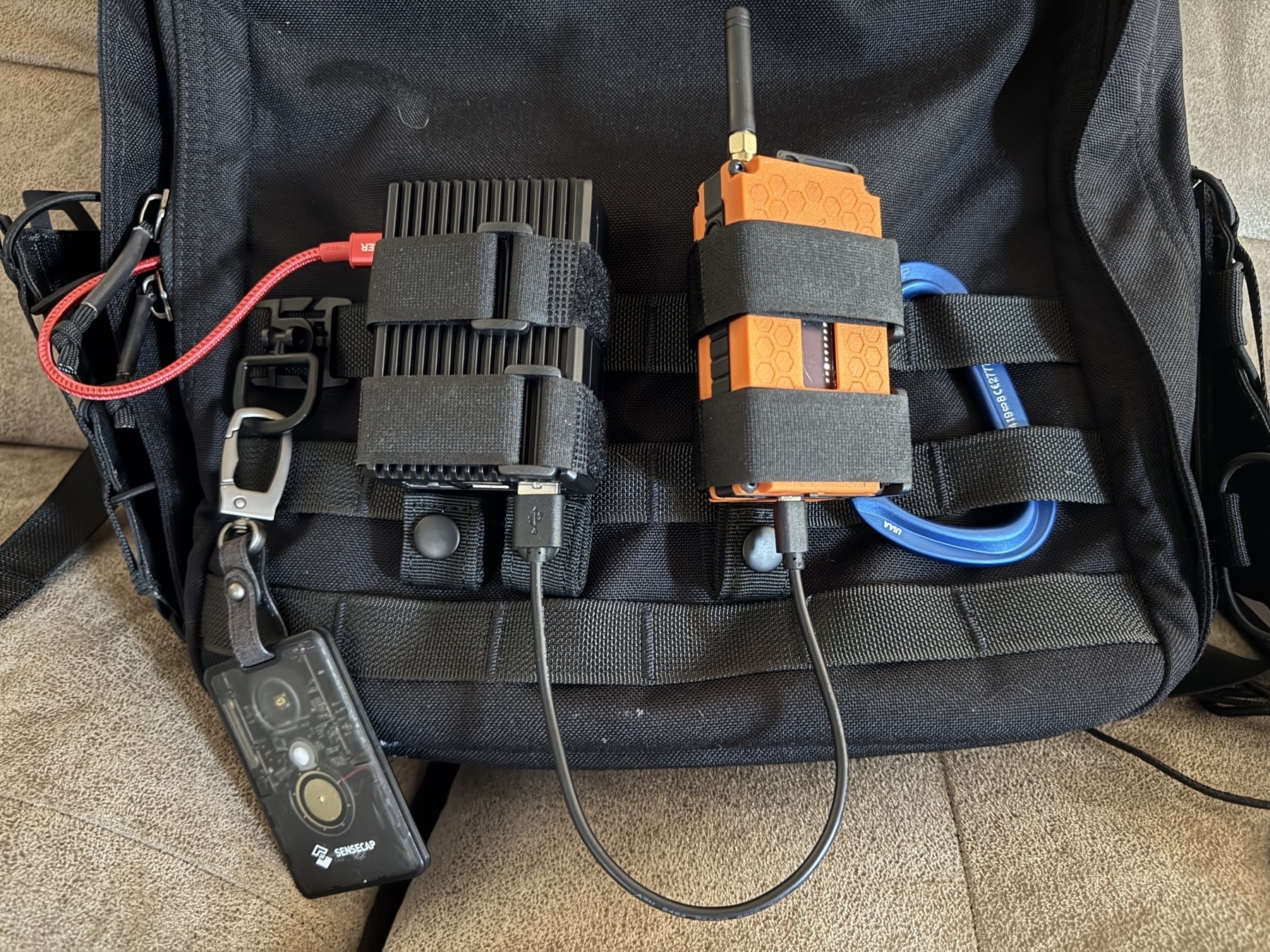 A backpack with MOLLE straps has a RAK 4631 mesh radio and a little Raspberry Pi case attached to it. There are power and connection USB cables dangling. A SenseCAP T1000-E is clipped on for the ride.