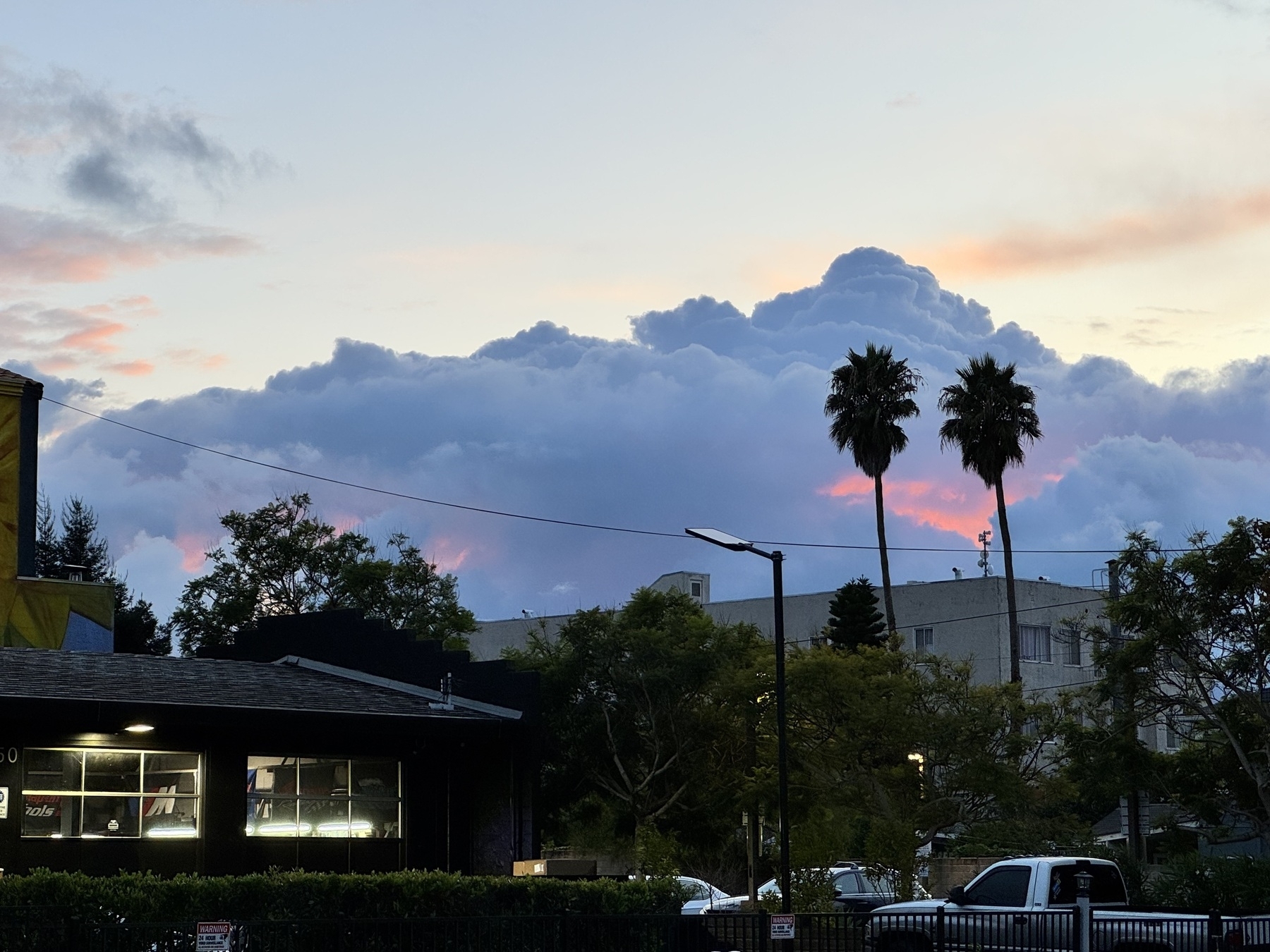 Giant blue-grey clouds behind a close skyline with palm trees. There’s a deep red glow inside several pockets within the clouds.