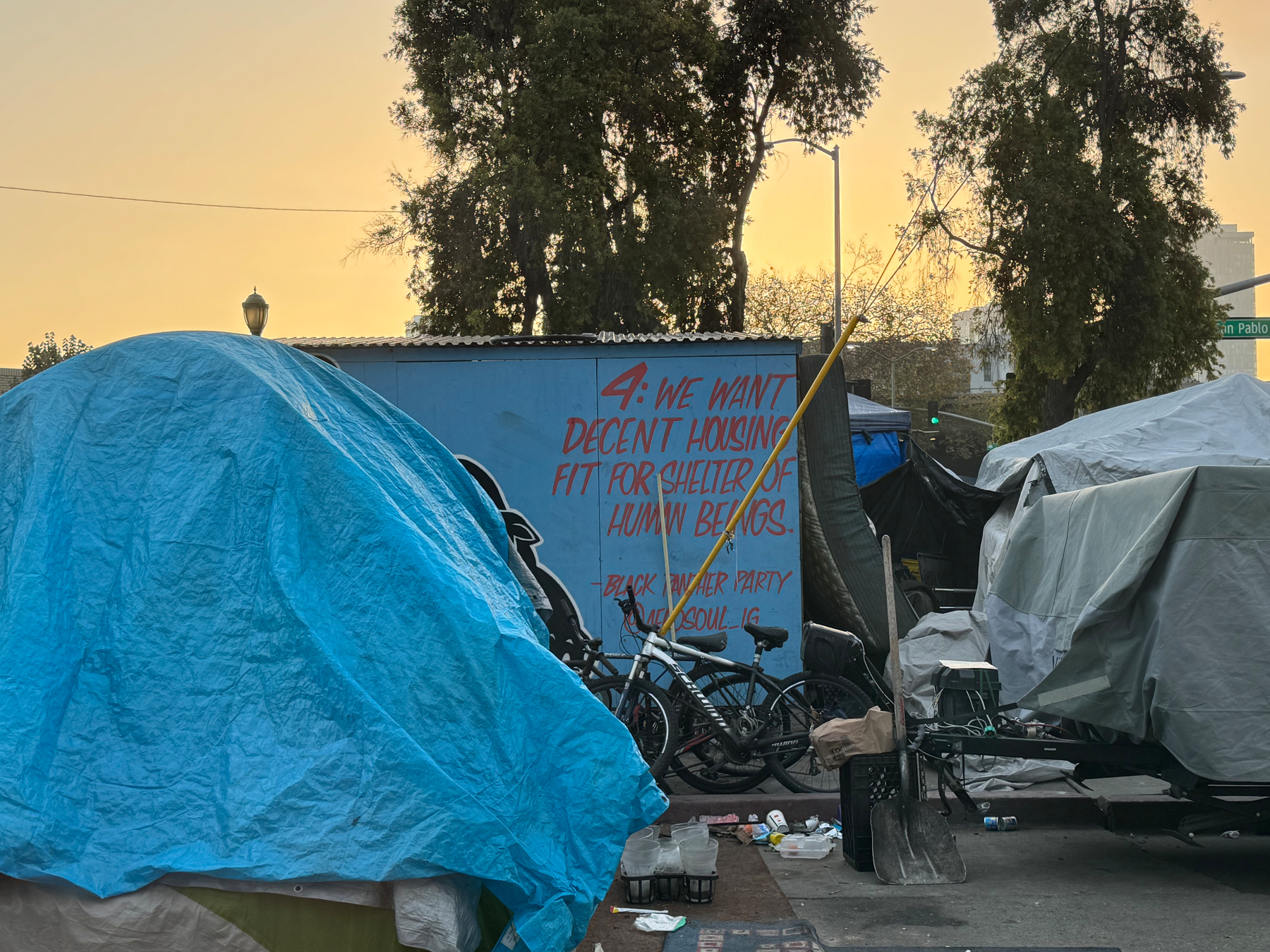 Photo of tents and bikes and trash under a yellow sky over a homeless camp. A sign behind it reads “4: We Want Decent Housing Fit For The Shelter of Human Beings.”