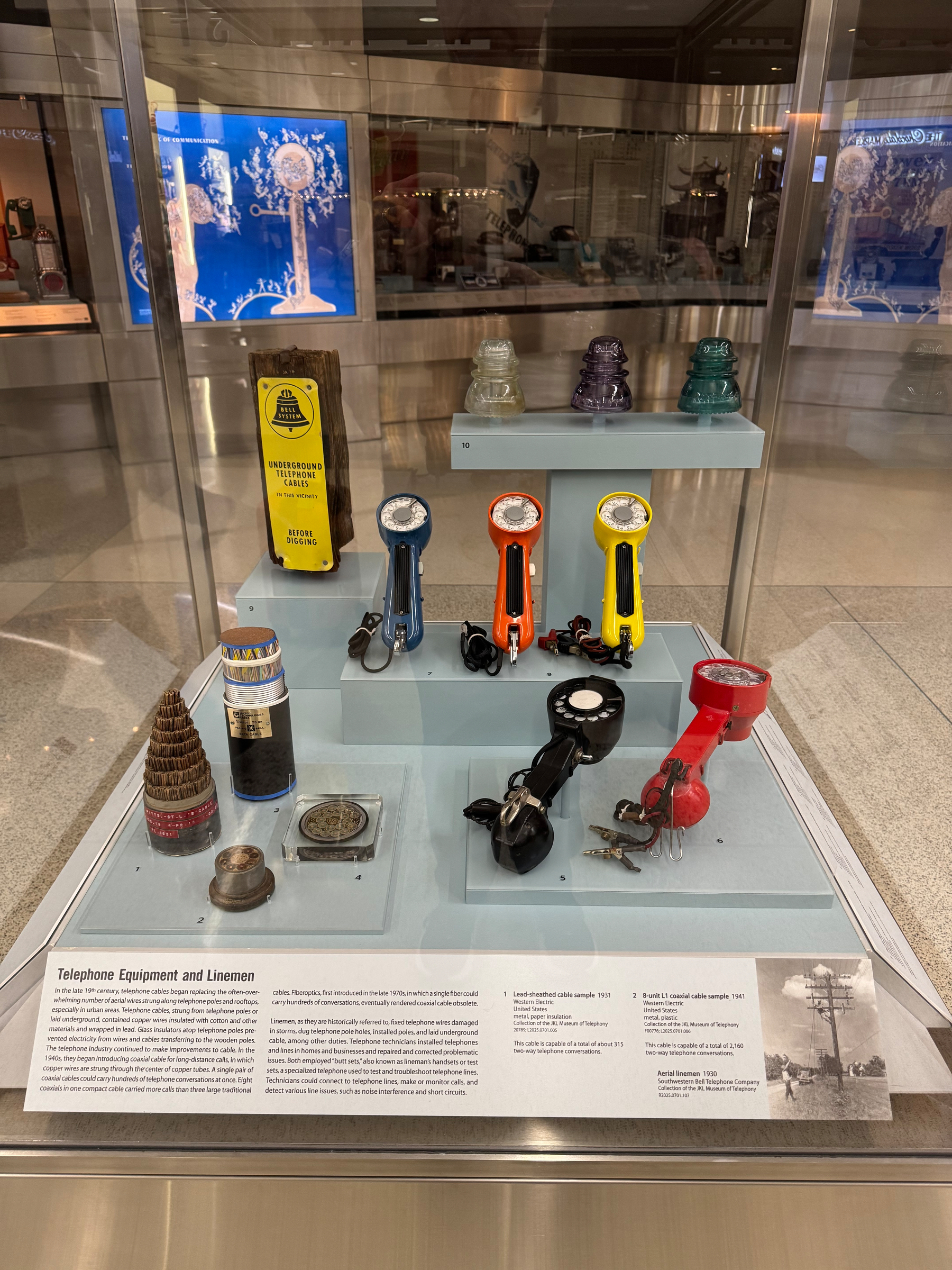 A glass museum case full of blue, orange, yellow, black and red lineman test sets, and thick, like 3 inch wide, cross sections of telephone cables.