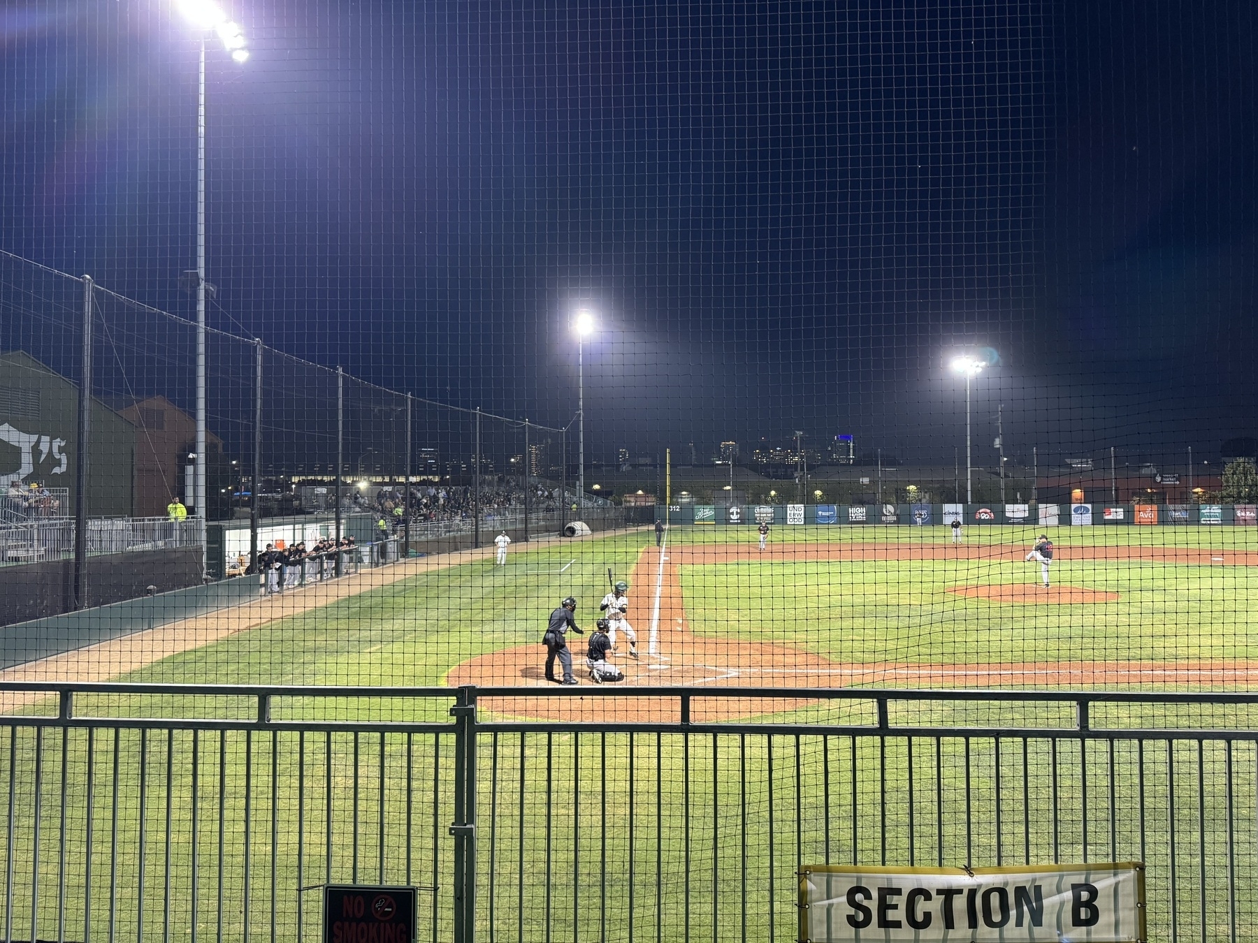 A baseball diamond taking from behind home plate, looking down the 3rd base line. The batter is set. The pitcher is winding up. It’s night time and the stadium lights are on. The glow of Oakland, California’s skyline is low in the background.
