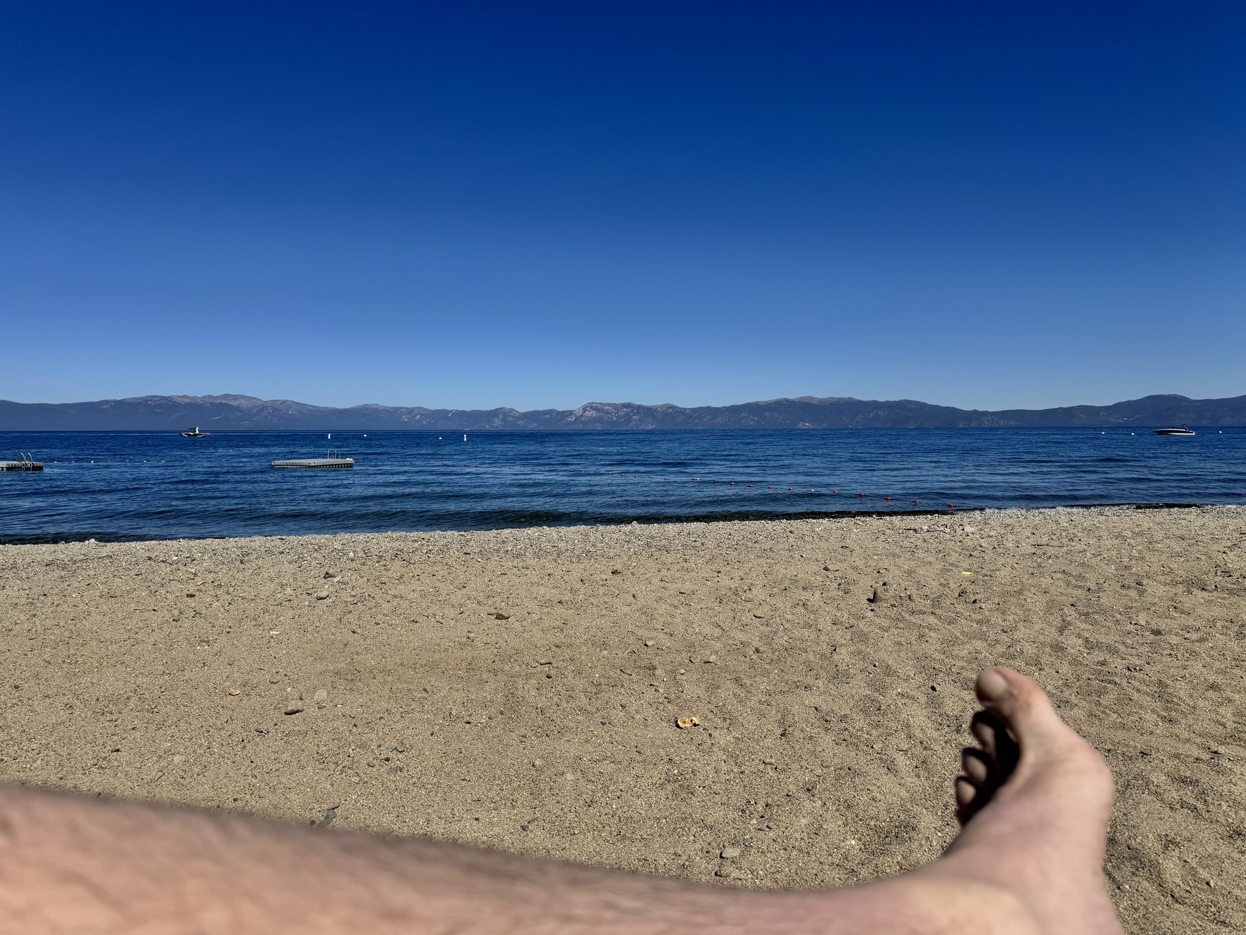 Photo of a beach with impossibly blue water and mountains in the distance. The edge of someone’s leg and foot is in the foreground.