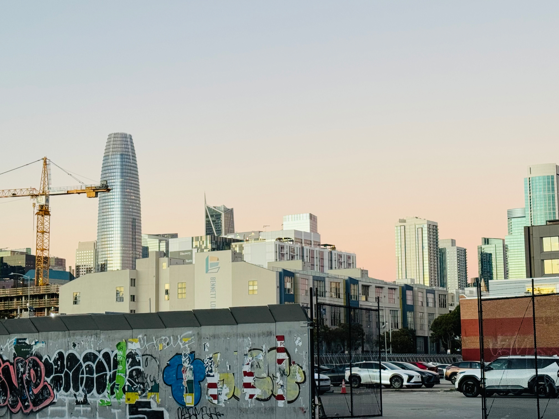 San Francisco from street level, looking up at tall buildings (including the Salesforce tower). The buildings are blue and shiny. The background is pastel pink. A low wall in the foreground is covered with graffiti.
