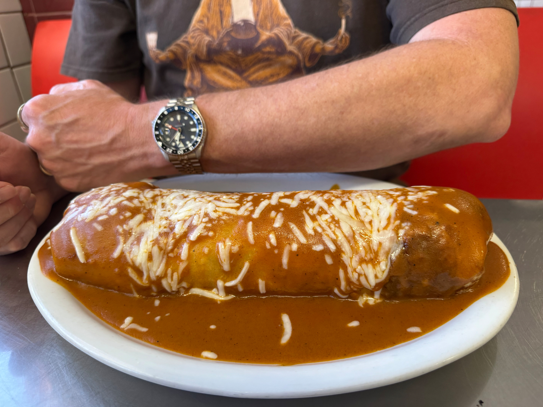 A plate-filling burrito is on a shiny steel table. Behind it is a man’s arm for scale. The burrito, or just plain burro, is the length of his elbow to wrist, but bigger around.