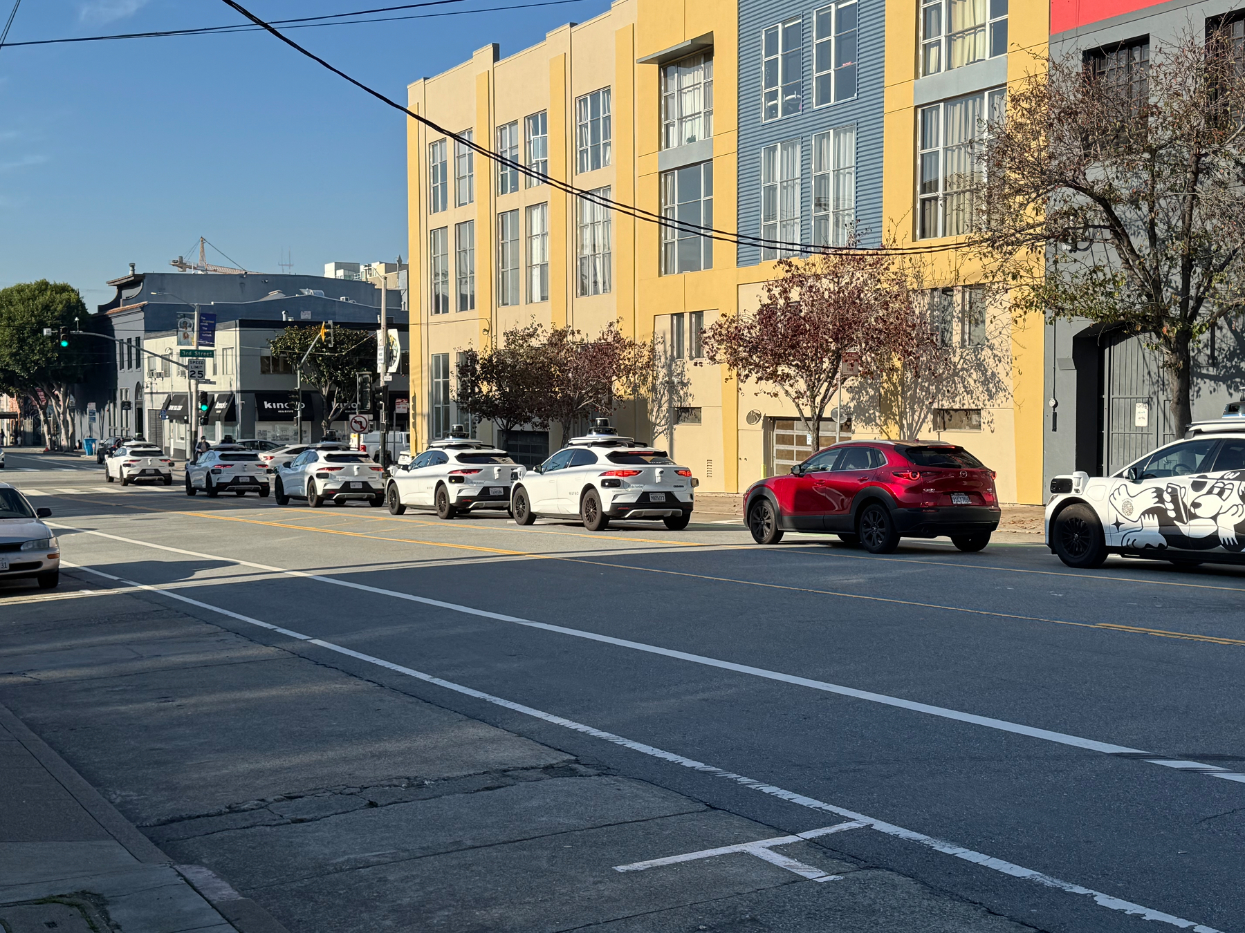 Photo of cars on a street moving toward a traffic light. There are 5 white self-driving Waymo taxis, a red car who wandered in, then another Waymo.