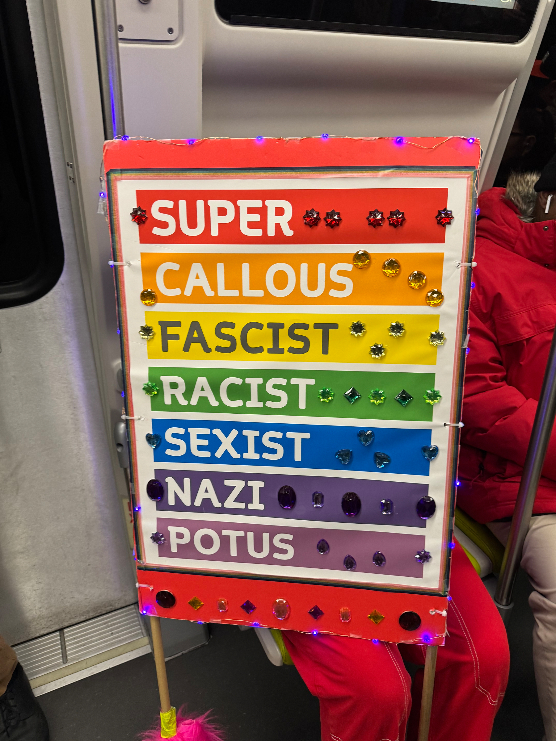 Picture of a rainbow colored protest sign someone’s carrying through the subway. It reads:&10;&10;SUPER&10;CALLOUS&10;FASCIST&10;RACIST&10;SEXIST&10;NAZI&10;POTUS