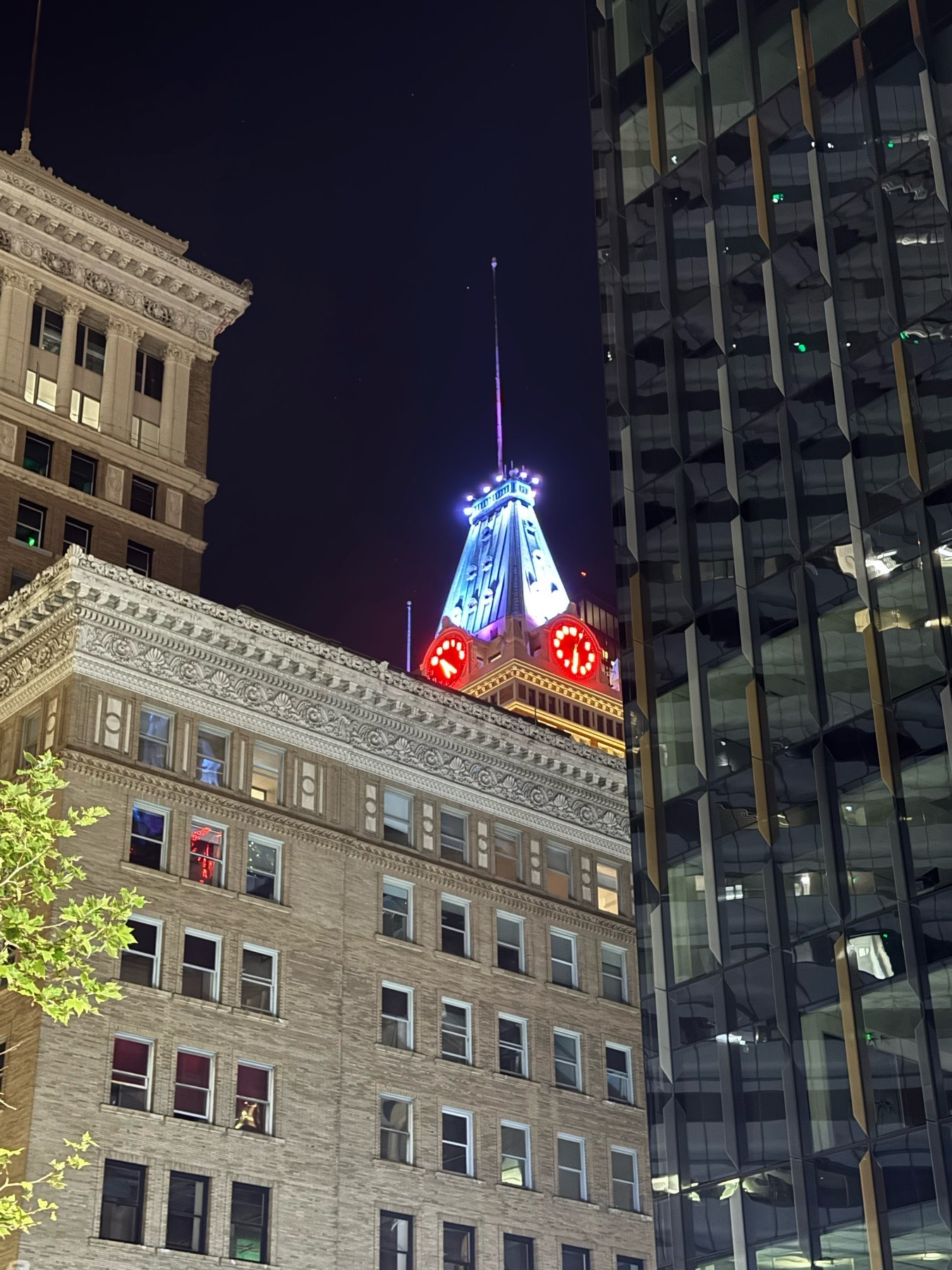 A brightly lit tower with neon lit clock faces rices above yellow brick and glass-and-steel office buildings in front of a dark night sky. 