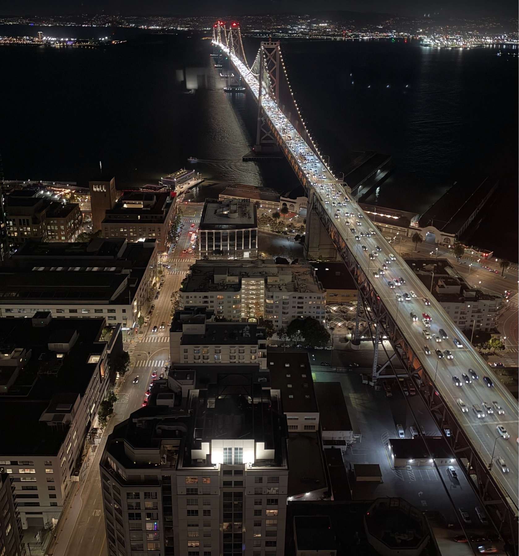 Photo looking down at the San Francisco Bay Bridge at night. There are a million headlights coming toward the camera. The bay is shiny and black under the clouded sky. Streets and buildings are lit with thousands of souls living their lives in the distance, alone, separate.