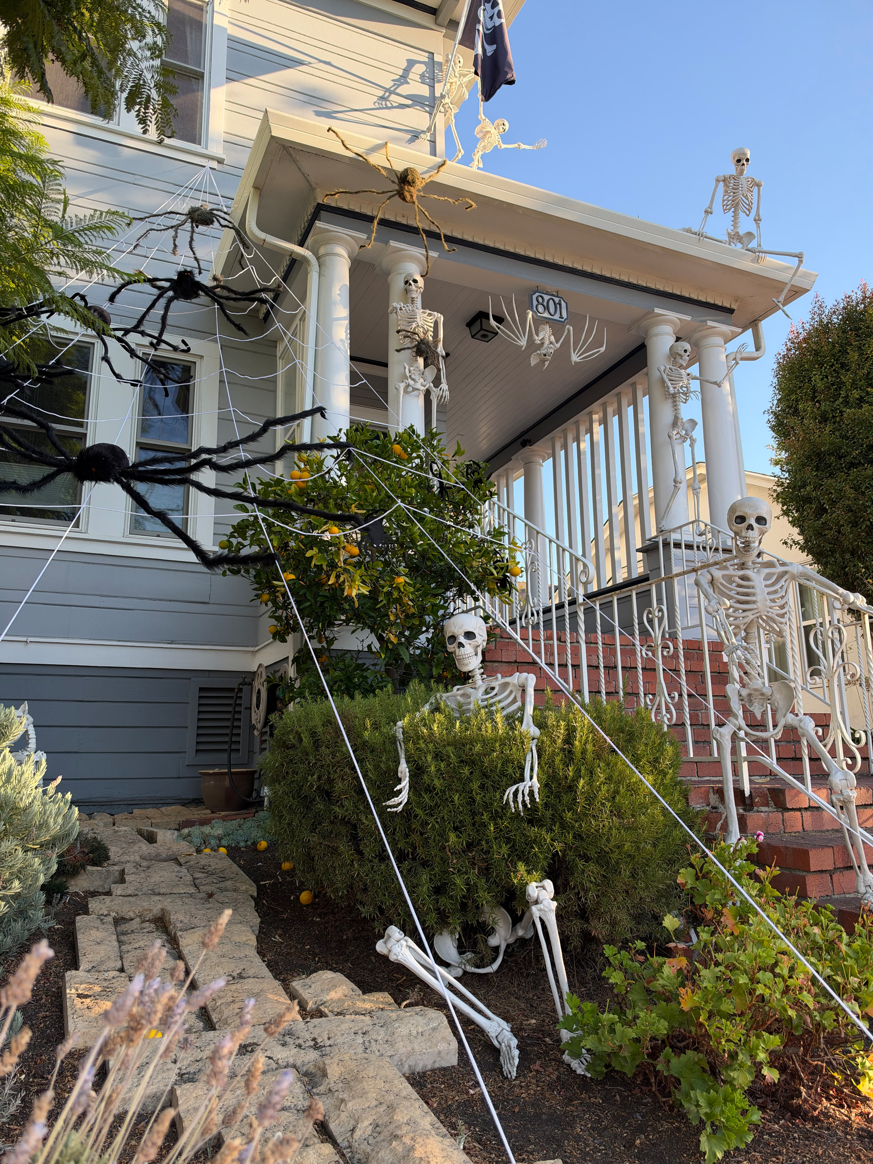 Photo of a Victorian house with spooky skeletons all over the place, in the gutters, peeking through shrubs, and hanging from columns. There’s a giant spiderweb with a 6 foot spider on it.