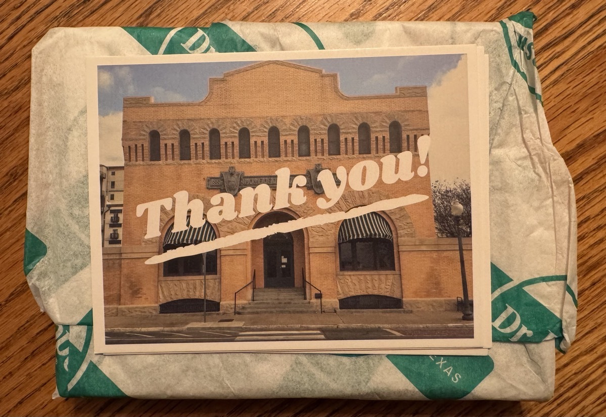A small package wrapped in paper, with a postcard of a southwest-style building sitting on it and covering most of it. The postcard says "Thank you!".