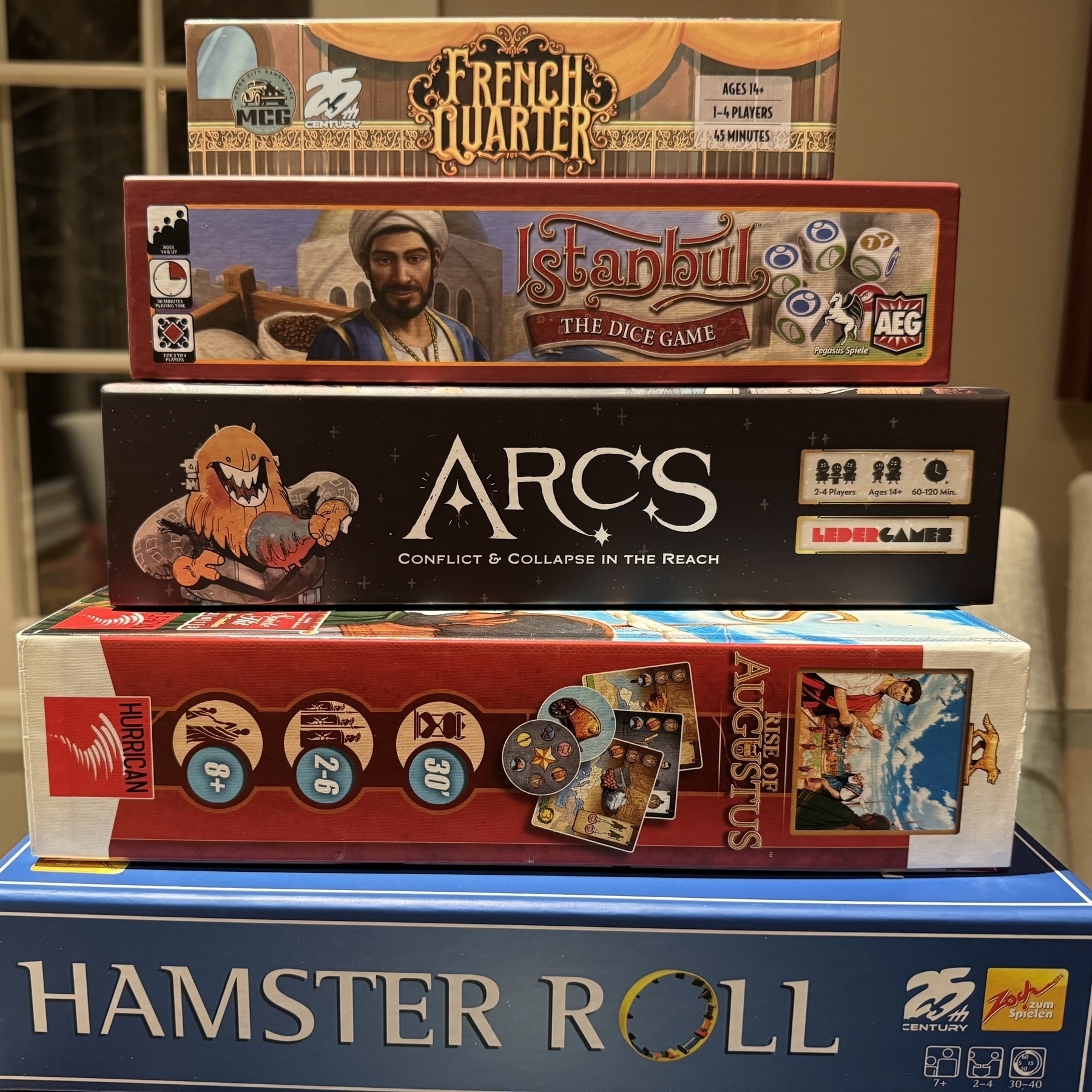 A stack of 5 board games. From top to bottom they are French Quarter, Istanbul the Dice Game, Arcs, Rise of Augustus, and Hamster Roll.