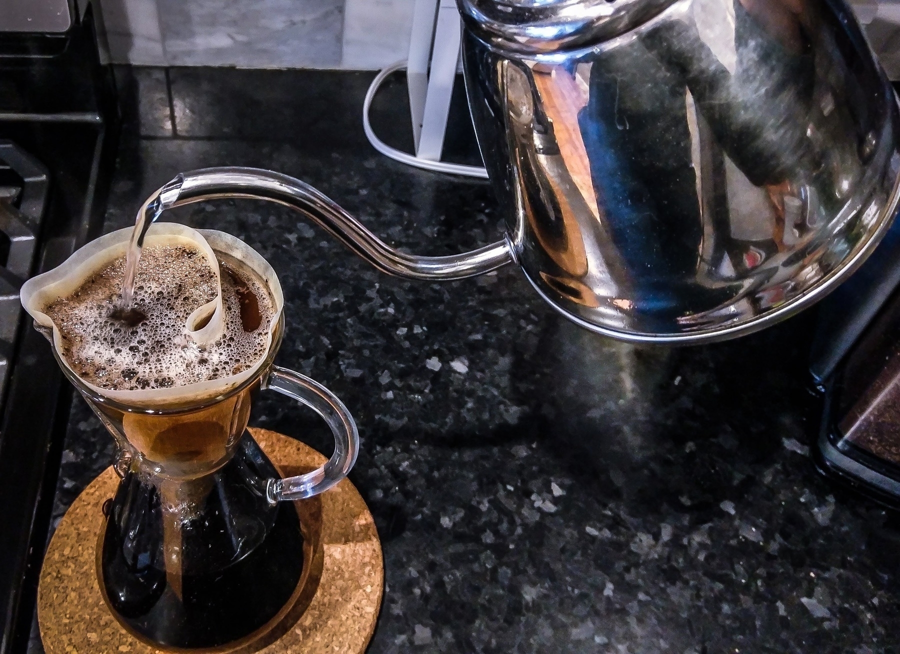 A chemex carafe of coffee (pour over), hot water being poured into it from a small stainless steel kettle. 