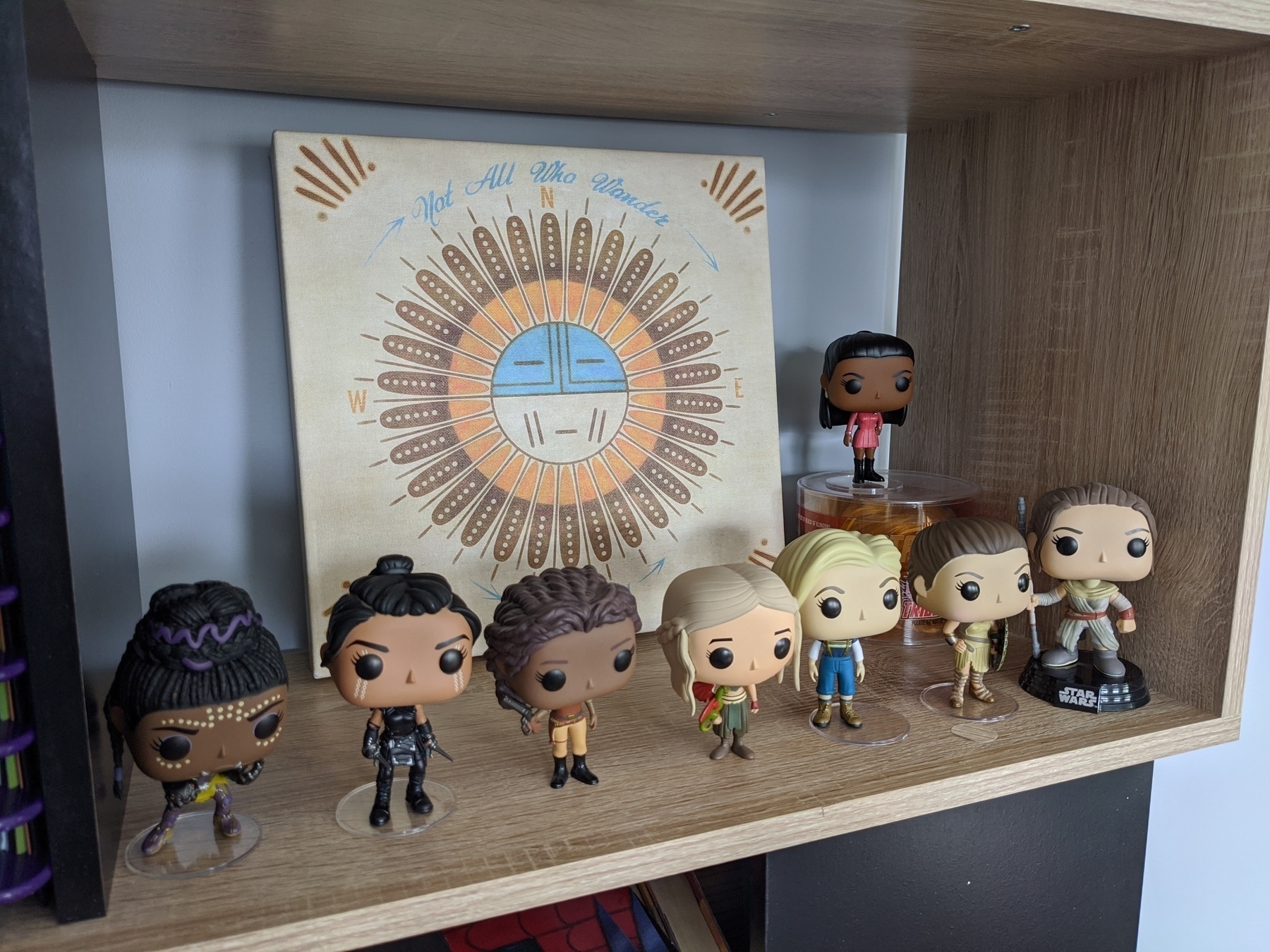 A light colored bookshelf covered in female (super) hero Funko Pops - Front Row, LtoR: Shuri, Valkyrie, Zoe Washburn, Daenerys Targaryen, the 13th Doctor, Wonder Woman, Rey.  Back Row: Lt. Nyota Uhura on top of a Tribble with a tiny Captain Kirk. There is a square canvas with a stylized sun and the phrase “Not all who wander are lost”.