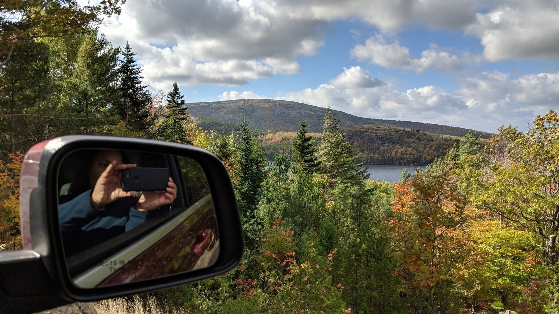A mountain landscape with a lake peeking through the trees. The photo is taken from a car and you can see the photographer and her cell phone in the car mirror. 