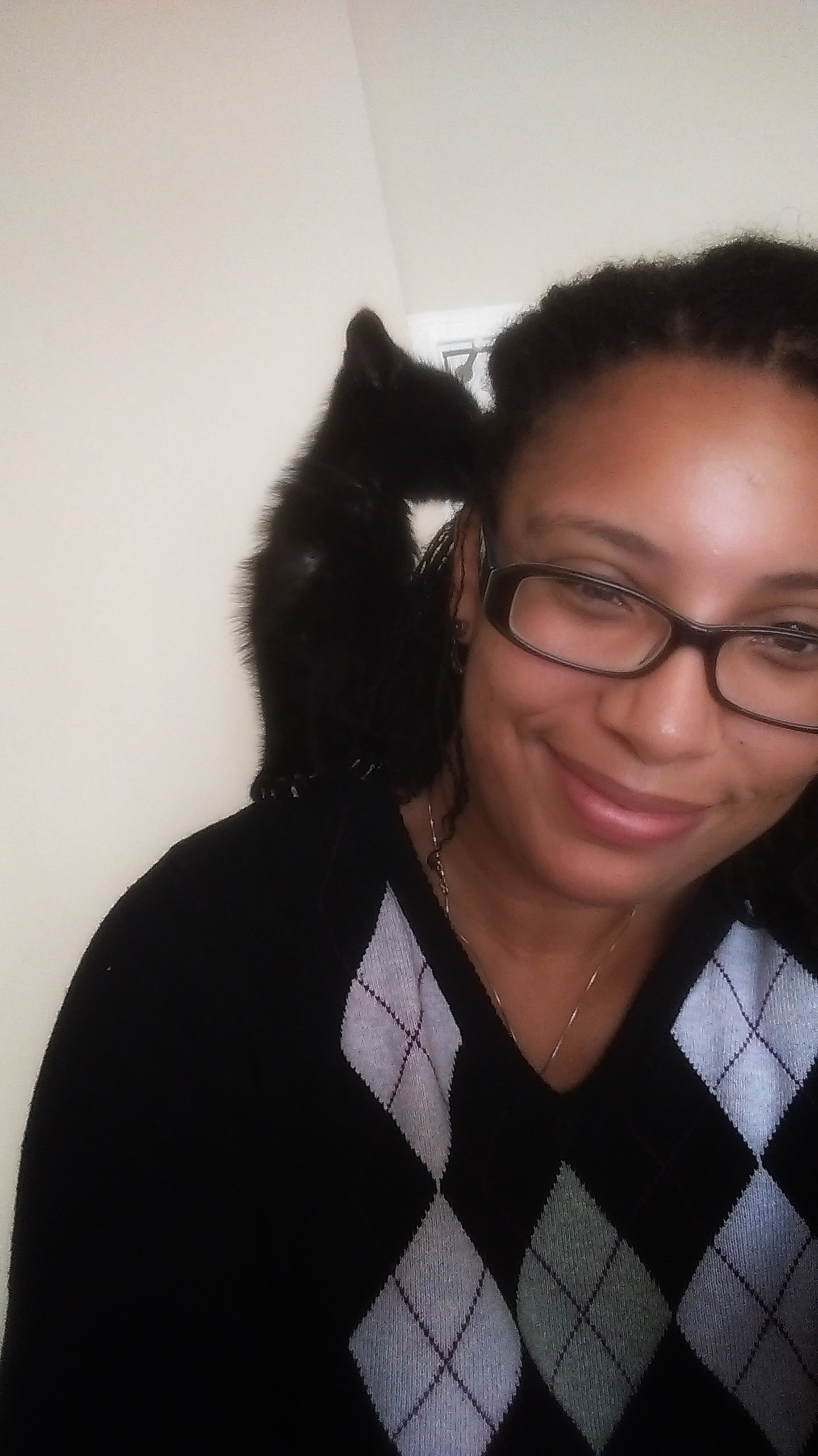 A tiny black kitten on the shoulder of a smiling brown woman. The kitten is licking her hair. 