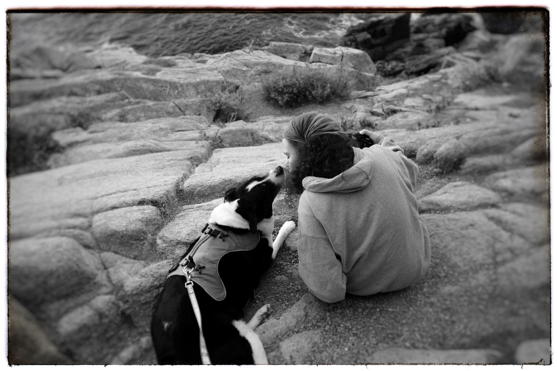 A black and white photo of a girl on the right appearing to kiss her black and white dog on the left. They are sitting on a rocky outcropping with waves crashing below.