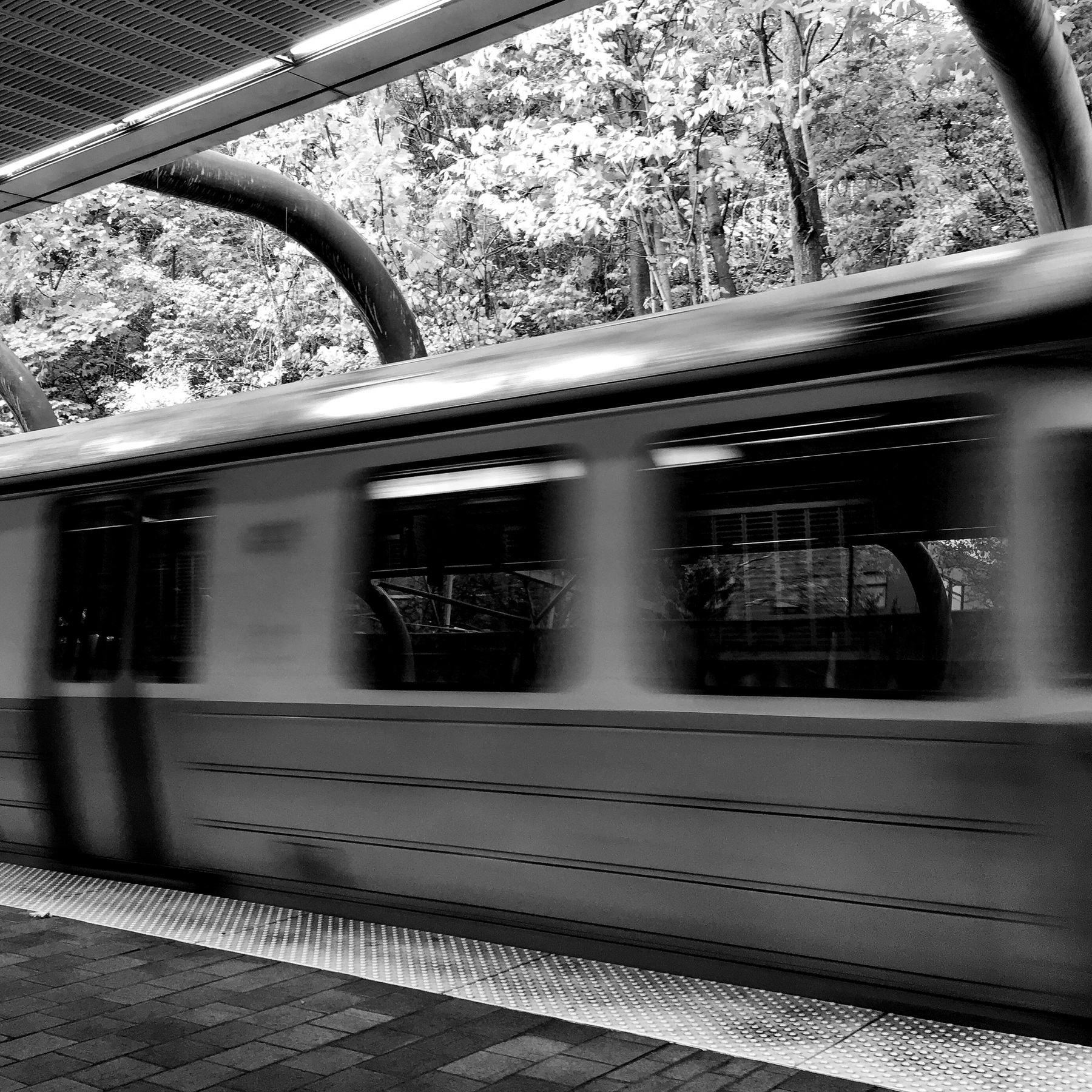 A black and white photo of a public transit train zipping by the platform. 
