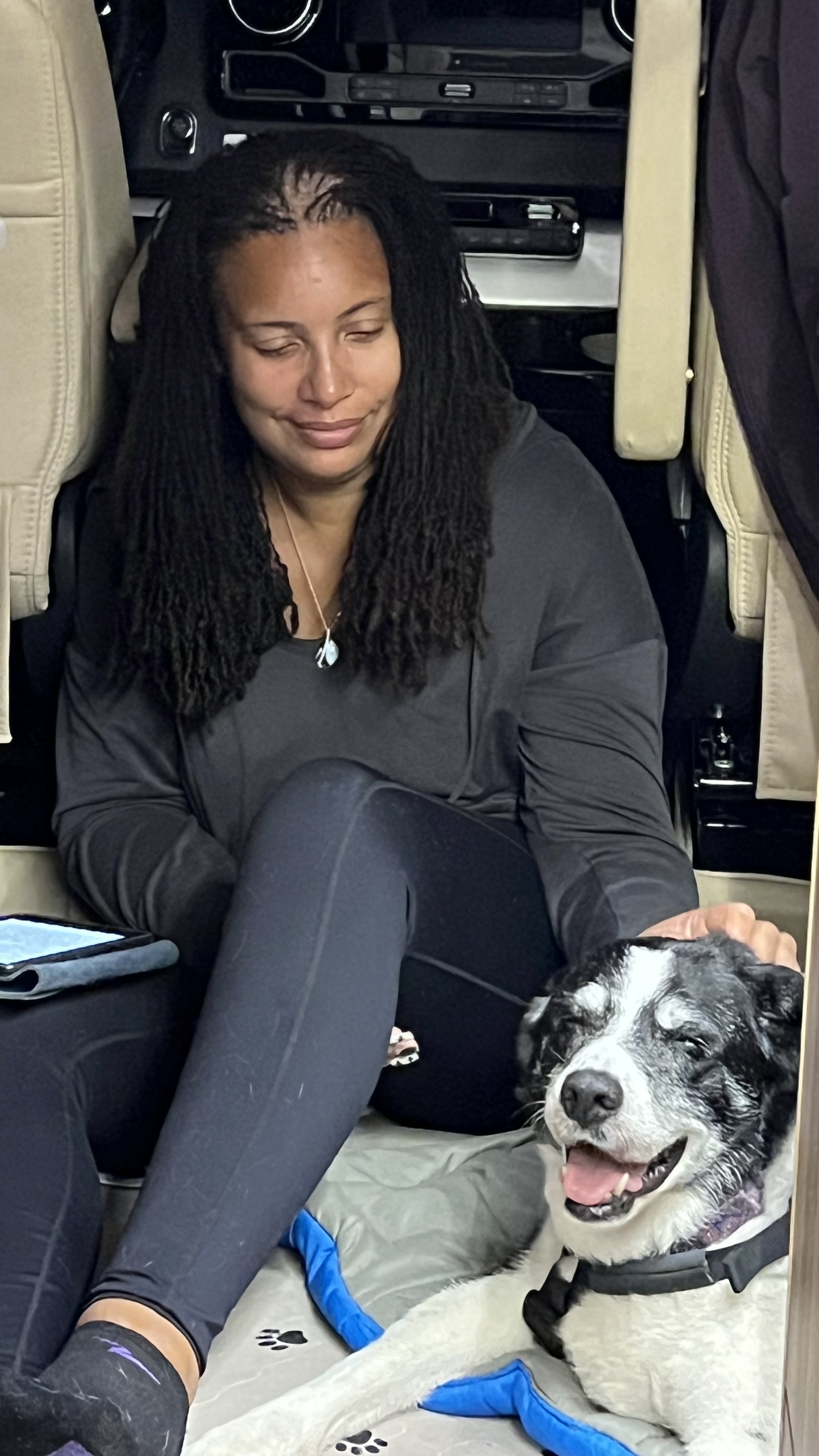 Brown woman with sisterlocks dressed in grey and black sitting next to and petting to a black and white border collie who appears to be smiling at the camera. 