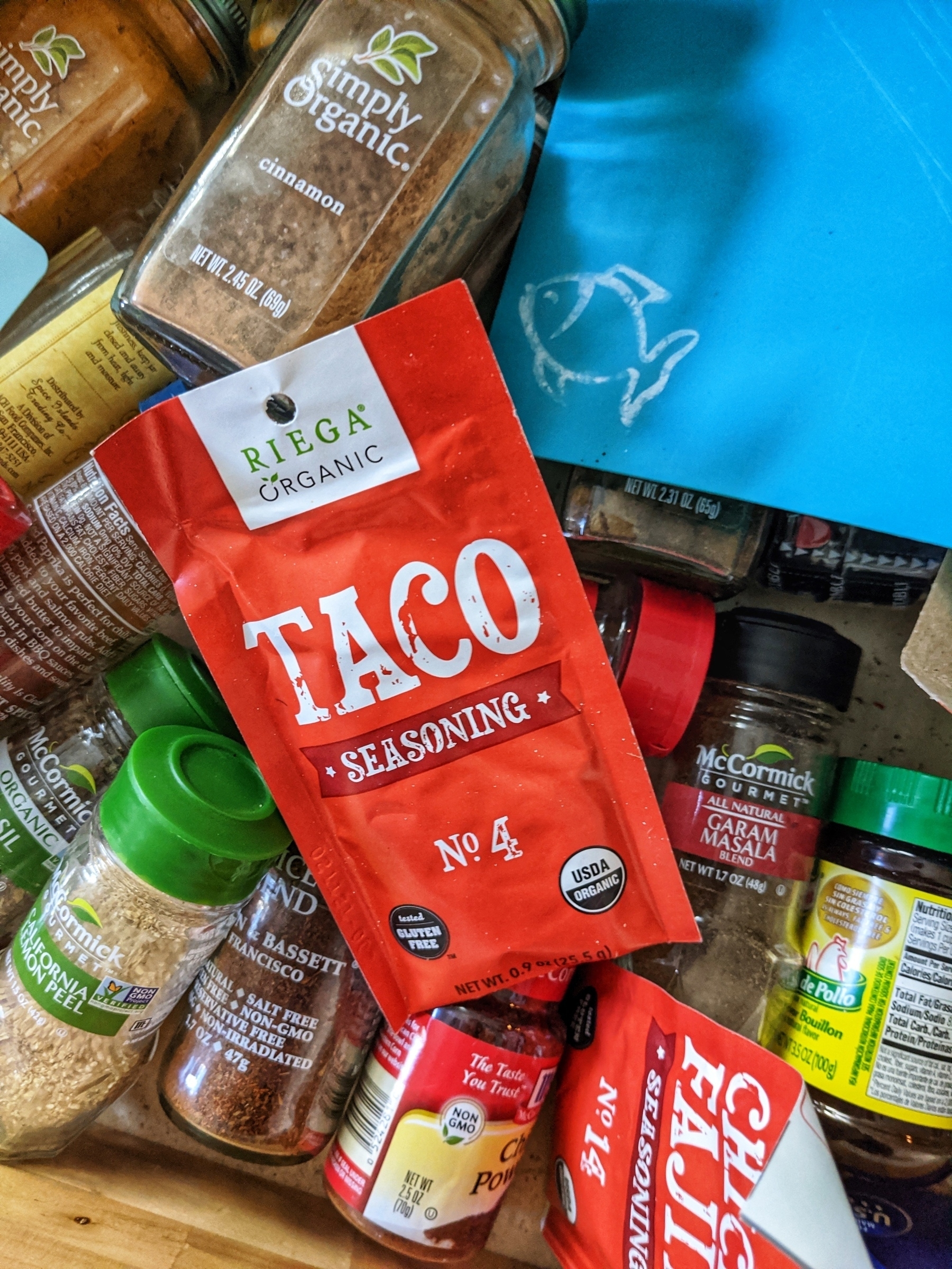 Jumbled spice jars in a drawer along with some taco and fajita seasoning packets and a plastic cutting board coded for fish. 