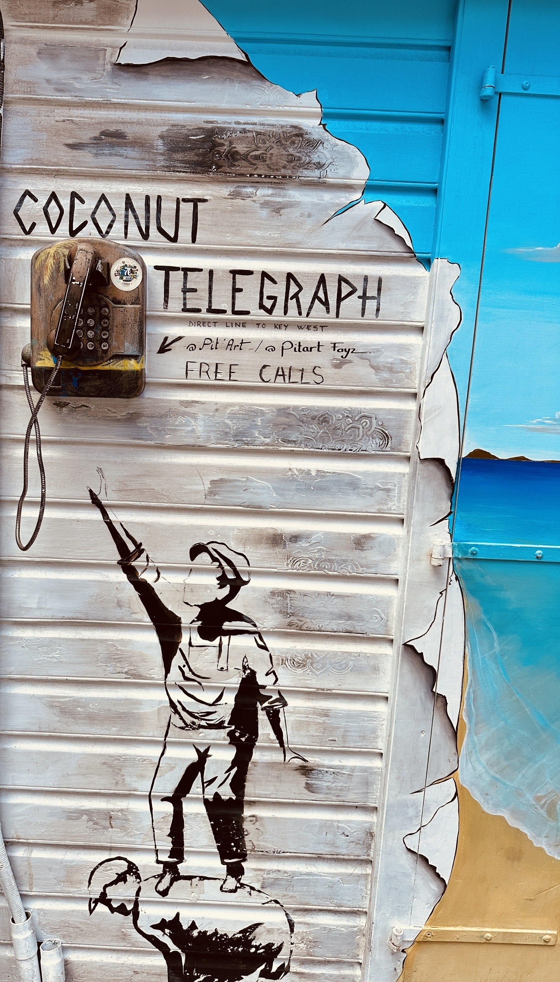Mural on a building in St. Bart’s: from the top “Coconut Telegraph” written above what looks like an old-fashioned pay phone with two male looking characters, one bending forward in order to be a lift for the second who is reaching for the phone. Additional text reads “direct line to Key West” and “Free Calls”. Below that, socials for @Pit’Art / @pitart foyez. 