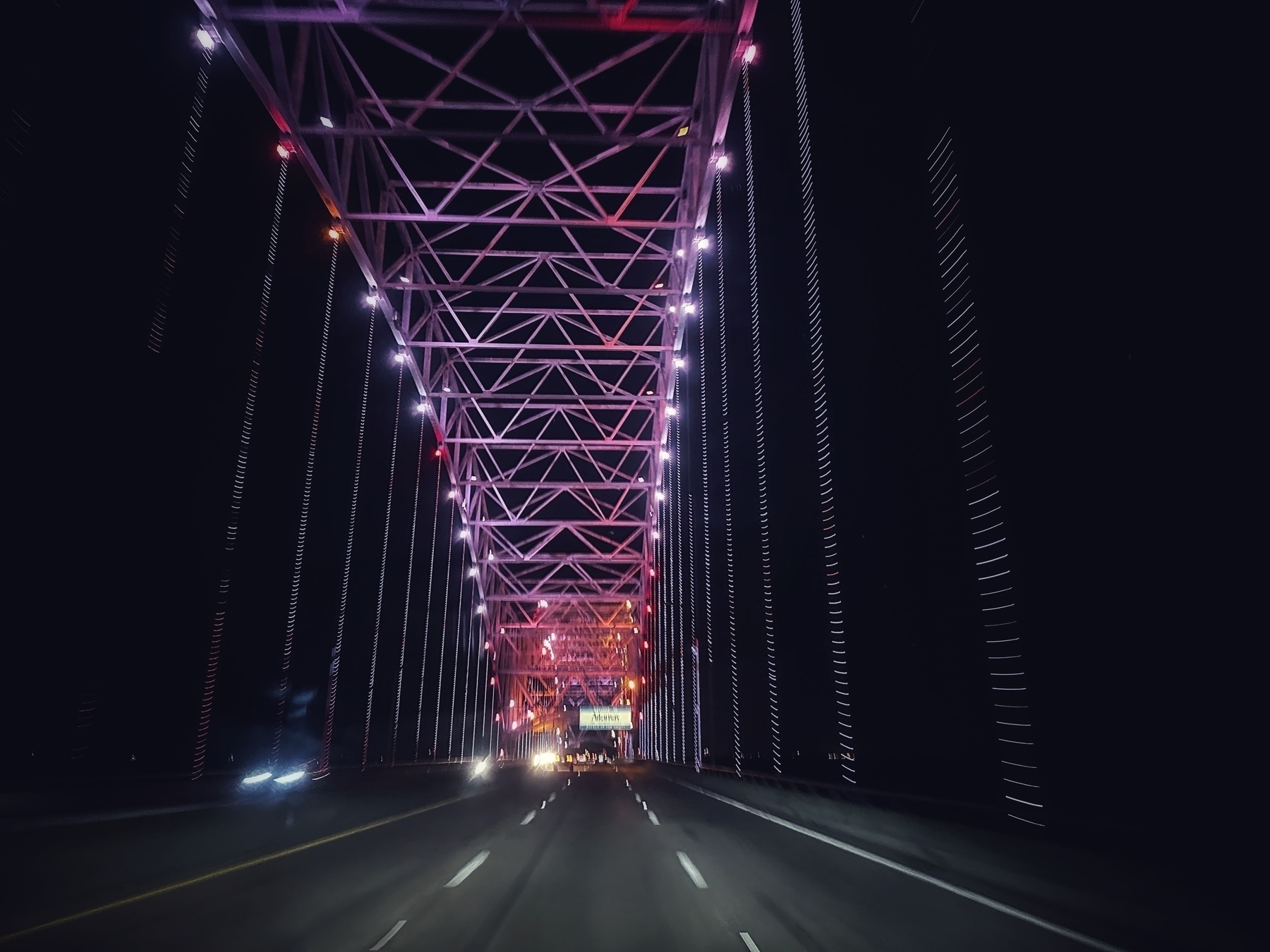 View of a bridge over the Mississippi River at night as someone drives over it. The bridge is lit up in shades of purple and pink with white pinpricks. The highway below is lit by the bridge, oncoming headlights, and the lights of the photographer’s vehicle. 