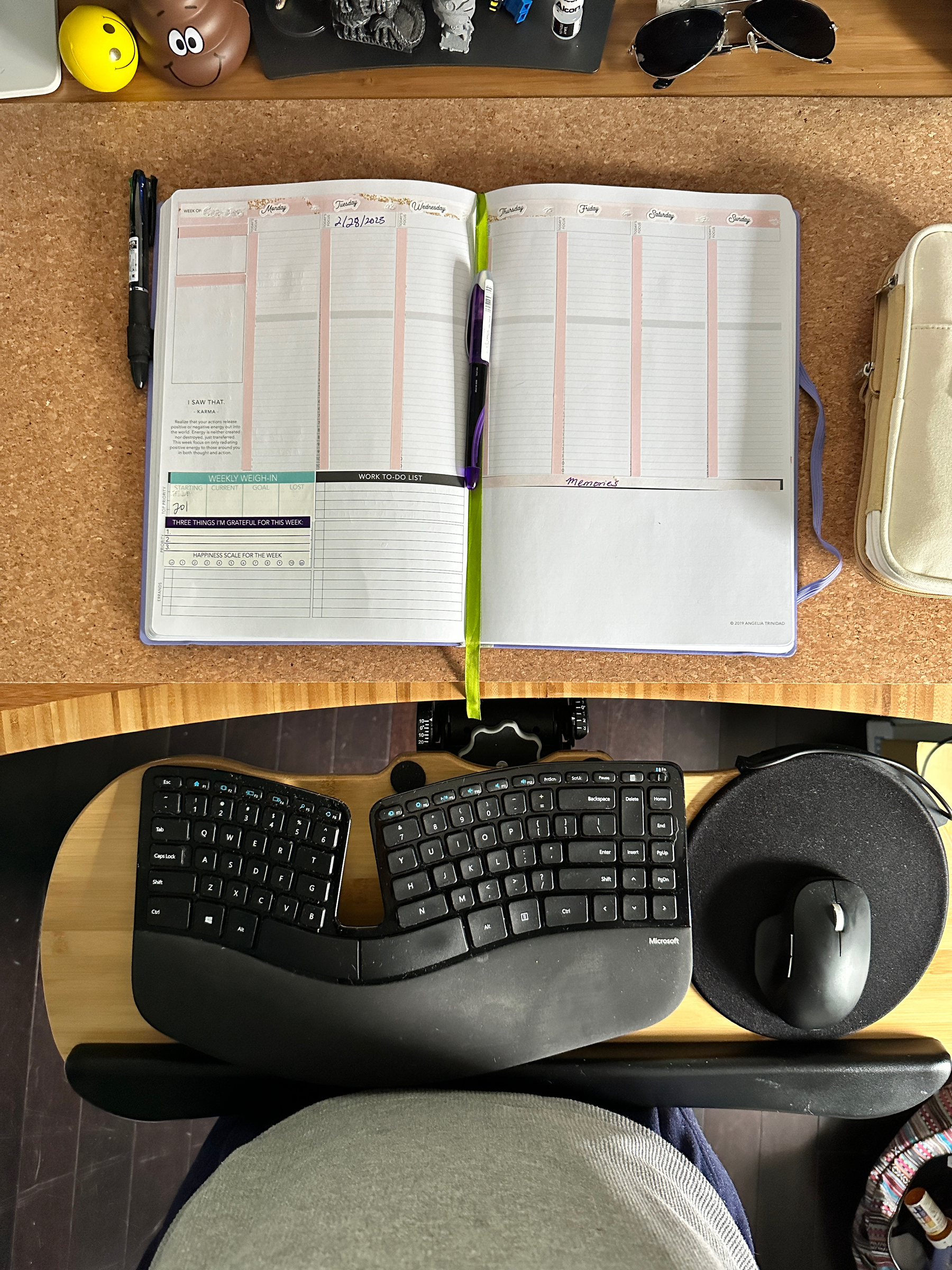 A top down view of a cork and bamboo standing desk. The author’s stomach can be seen in front of the split keyboard and mouse on a pullout tray. Above that is an open planner with a pen to the left and one in the center, a pencil bag to the right. Above that, various desk knickknacks including a shit emoji stress ball. 