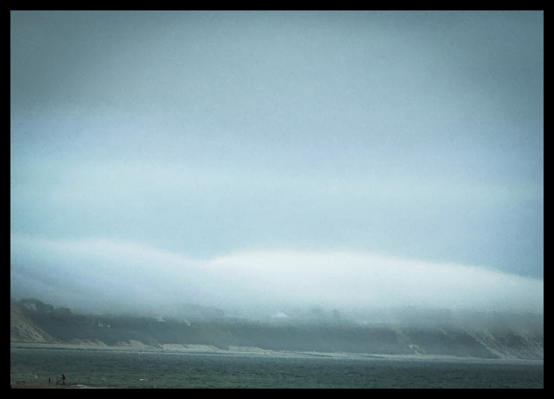 A seaside photo of the fog rolling over coastal hills and meeting the ocean. 