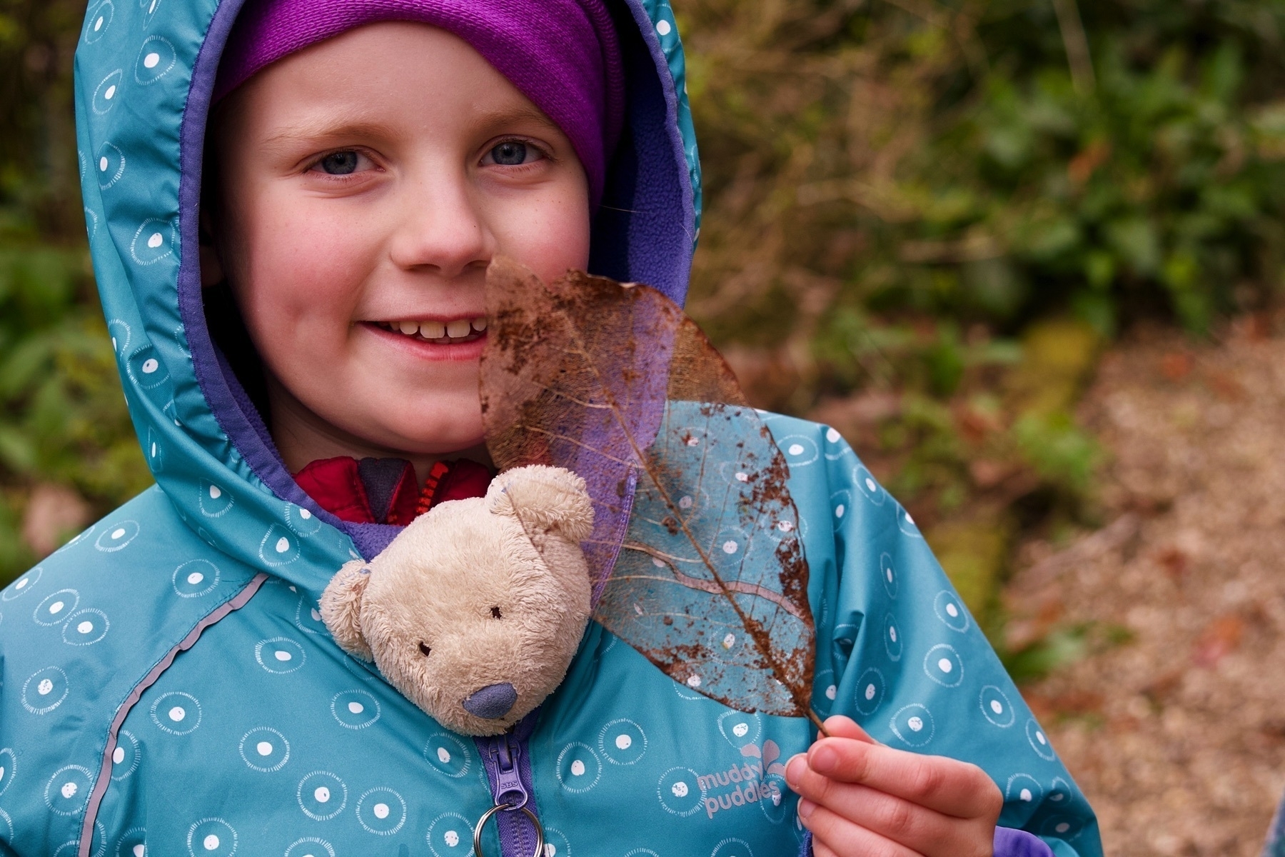 A girl with a teddy tucked into her coat holding a leaf skeleton. 