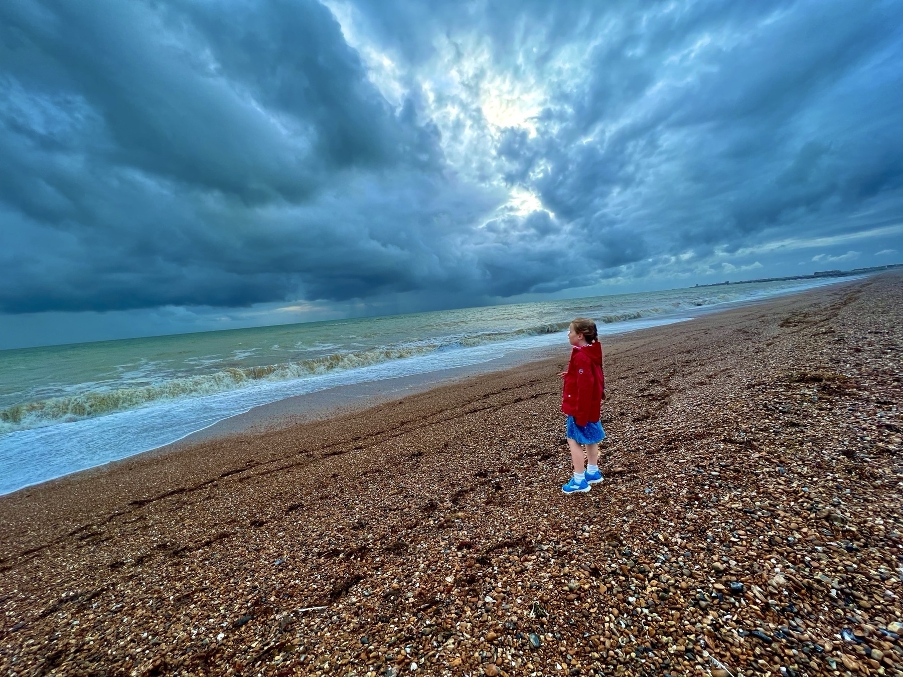 A girl standing on a shingle beach with stormy clouds overhead. 