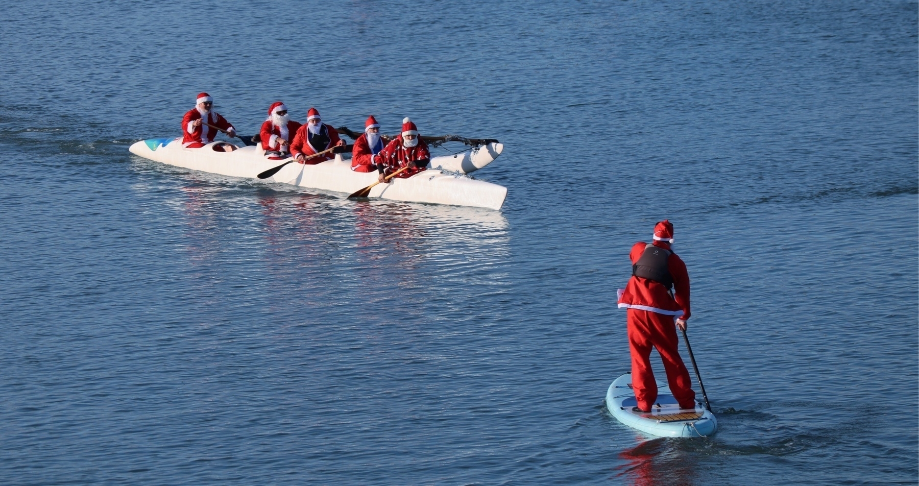 A santa on a SUP approaches santas in a boat. 