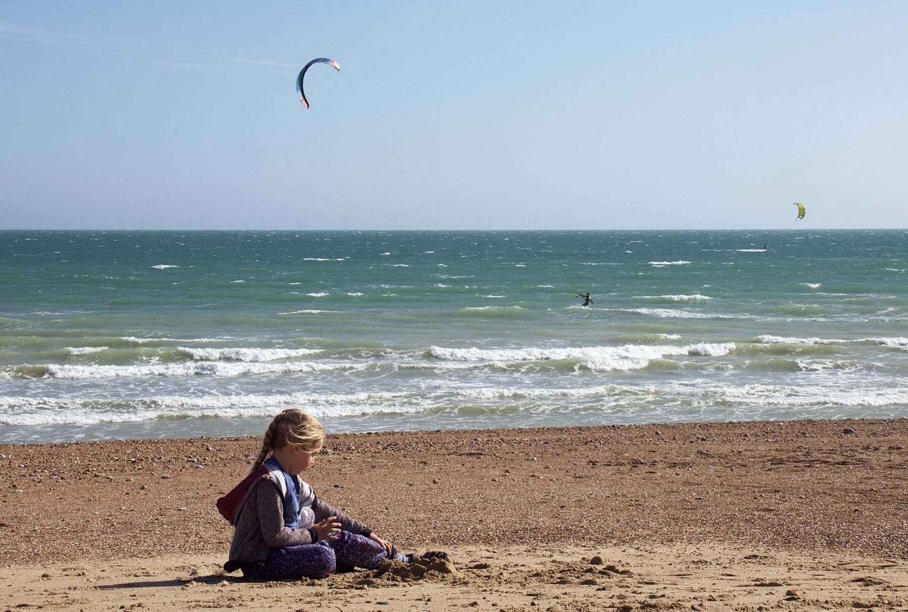A girl plays on the sand, with a kite surfer in the distance. 