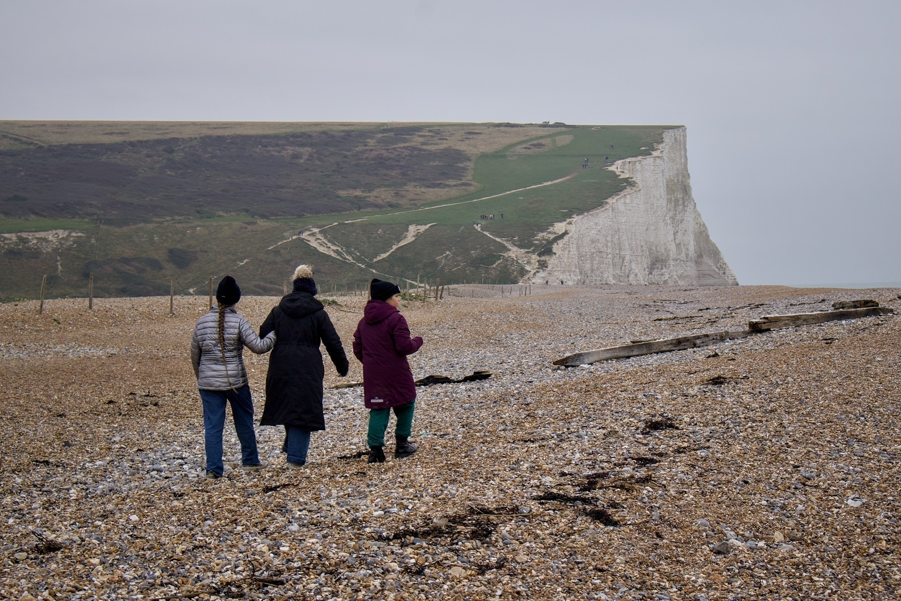 Three people walk along a pebble beach towards white chalk cliffs under a cloudy sky.