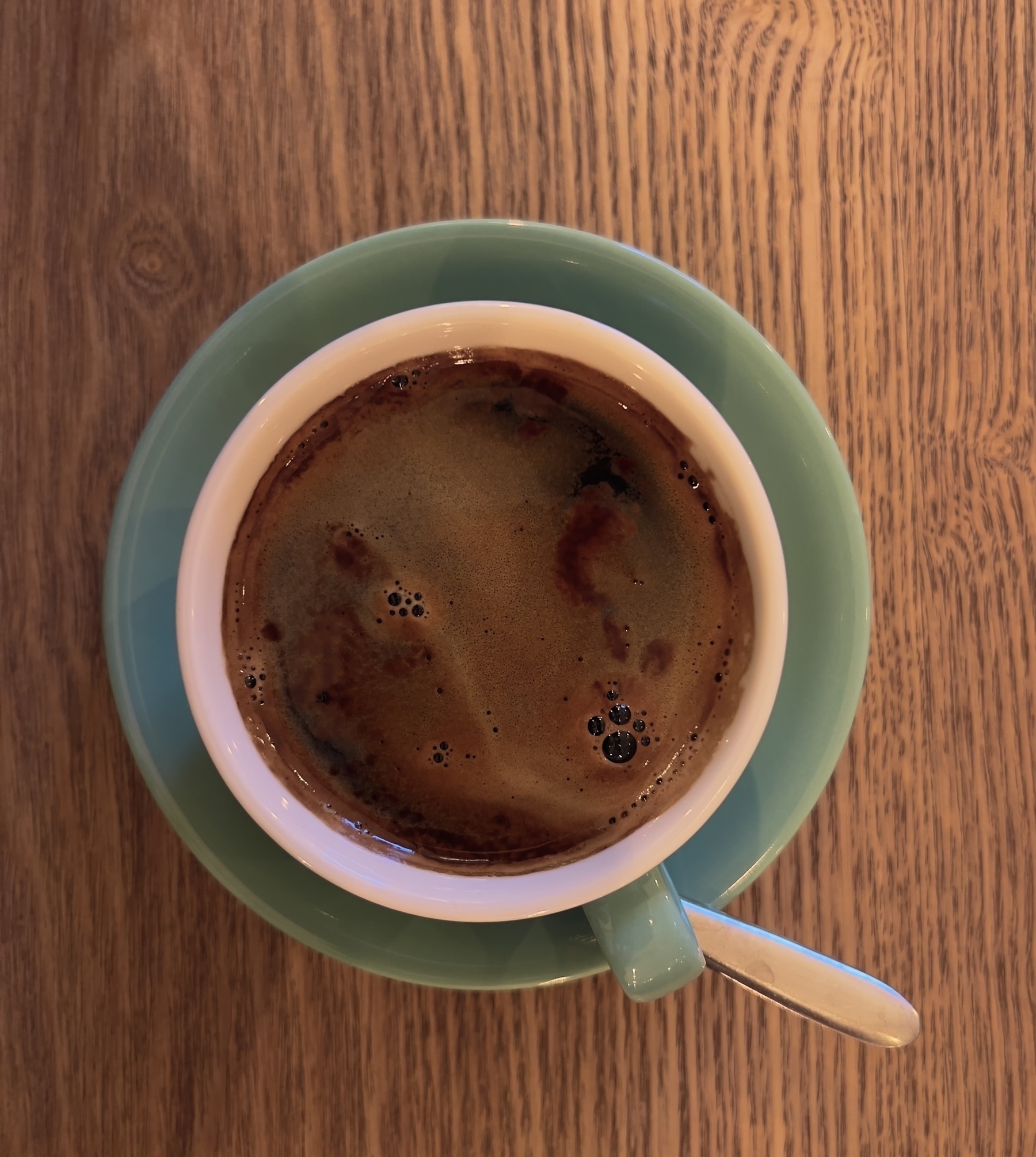 A cup of coffee with a frothy surface sits on a mint-coloured saucer on a wooden table.