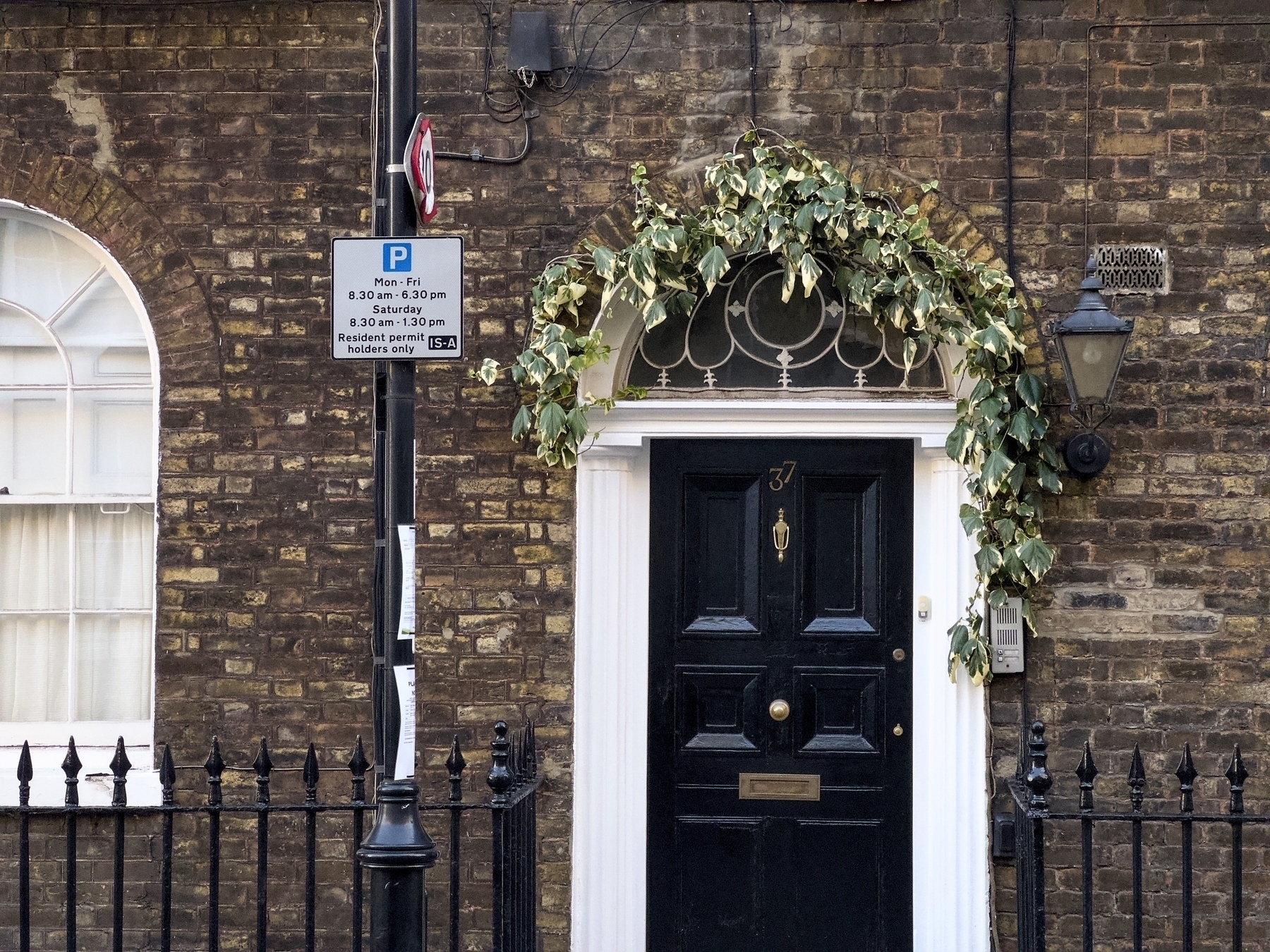 A black door with a decorative ivy arch is set in a brick wall, next to a parking sign and a traditional wall lamp.