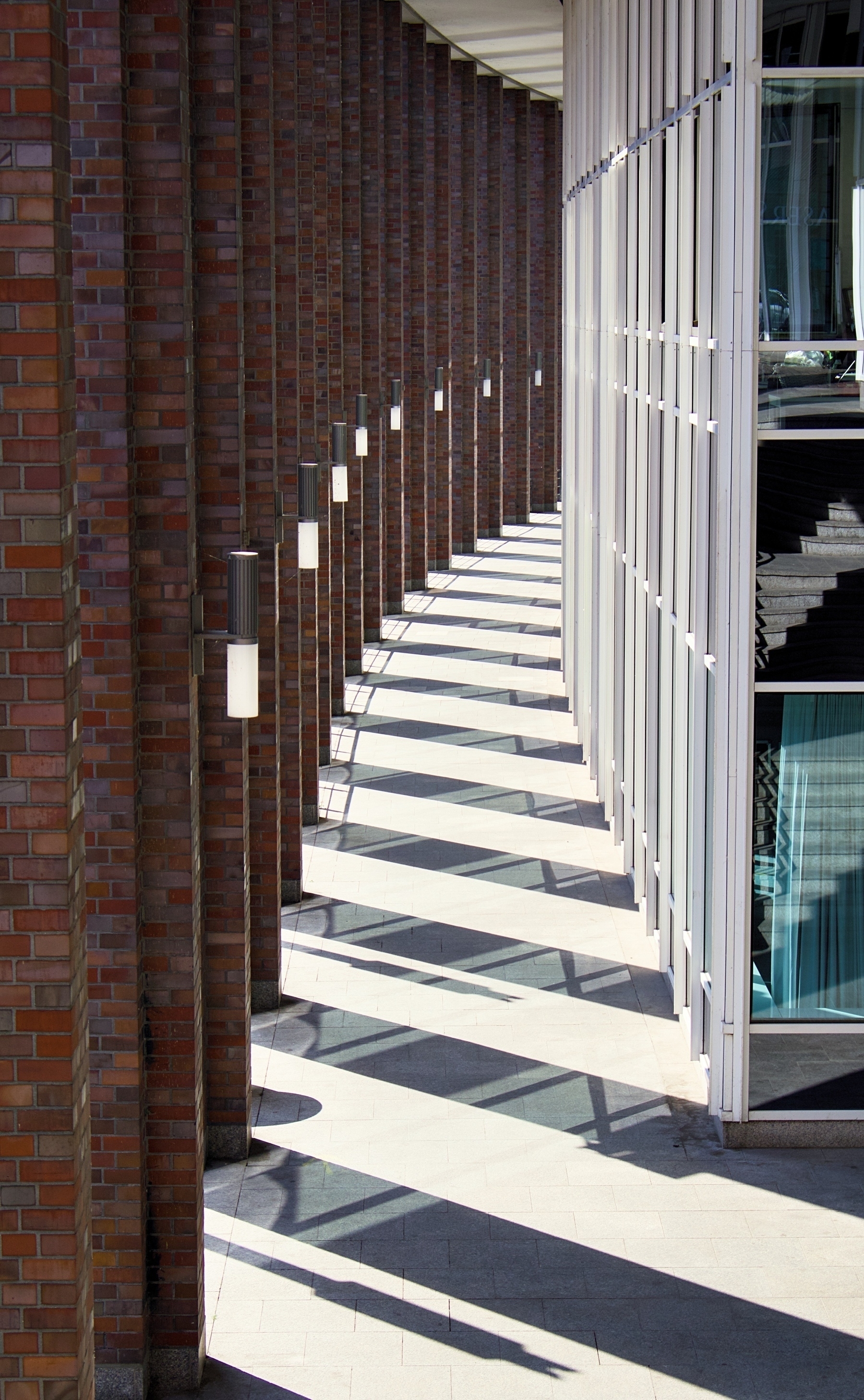 A corridor is formed by a curved brick wall with vertical white beams casting long shadows on the ground.