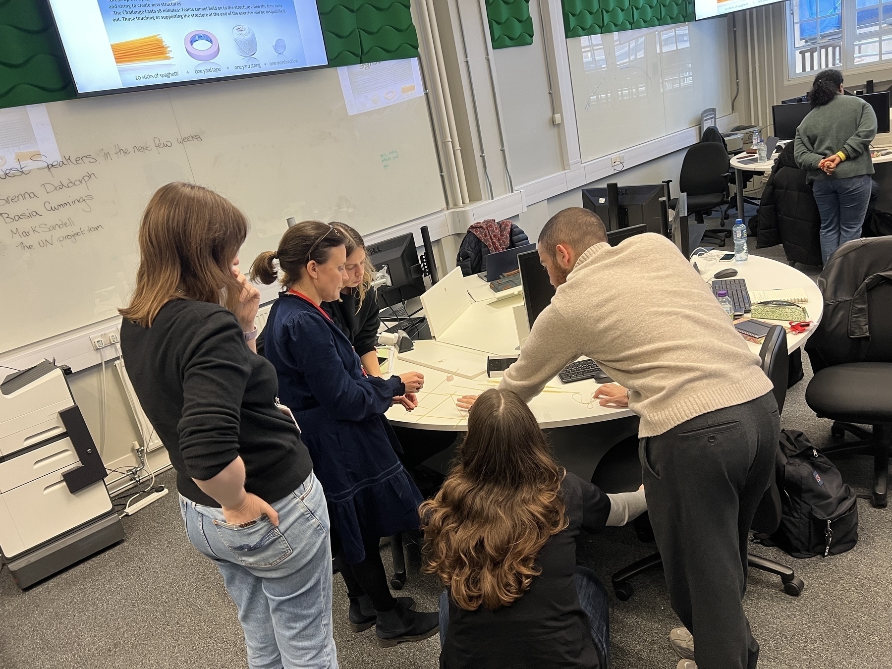 A group of people are gathered around a table in a classroom or office setting, engaging in discussion or work.