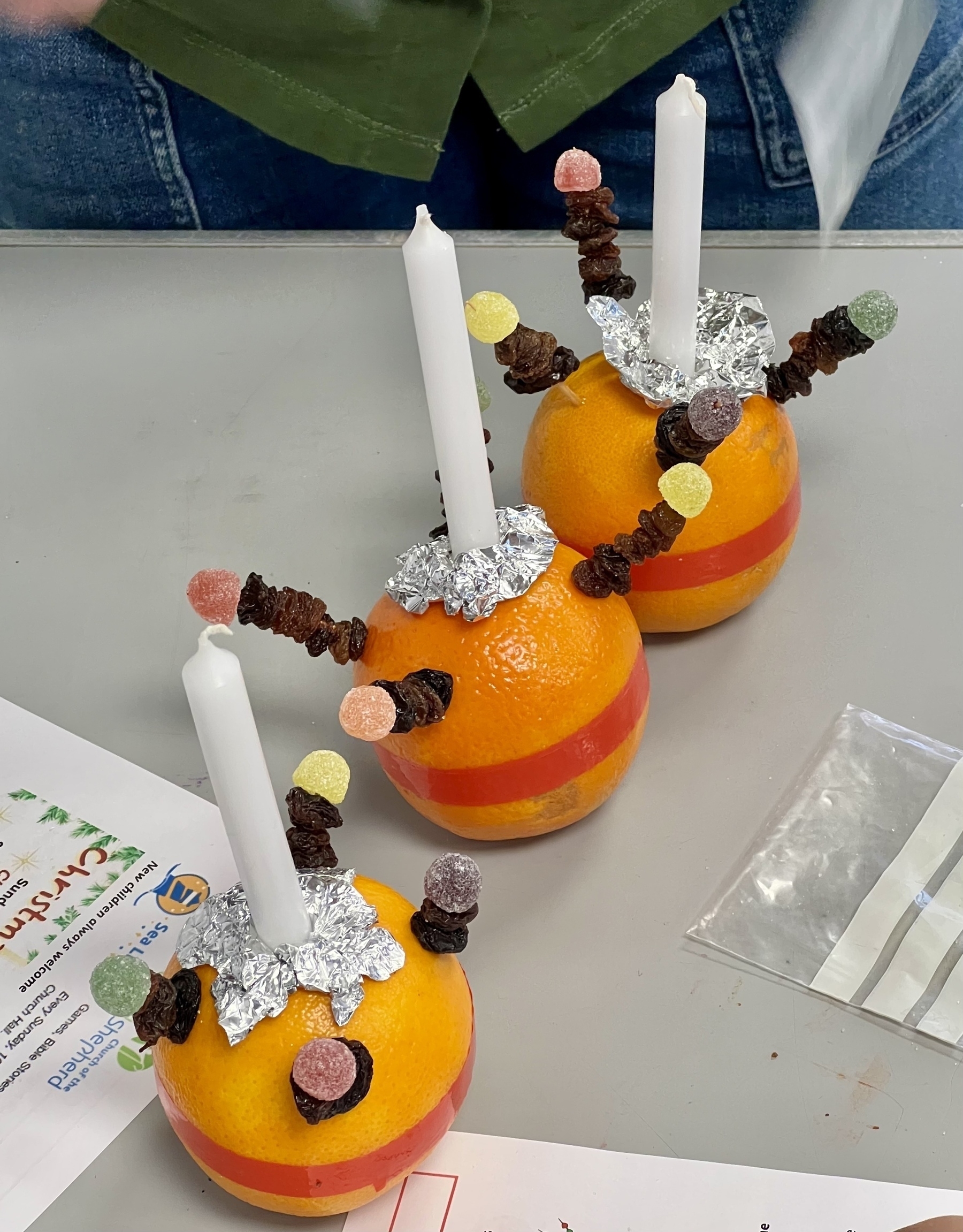 Three decorated oranges with candles on top, wrapped in foil, and adorned with gummy candies and dried fruit skewered on sticks.