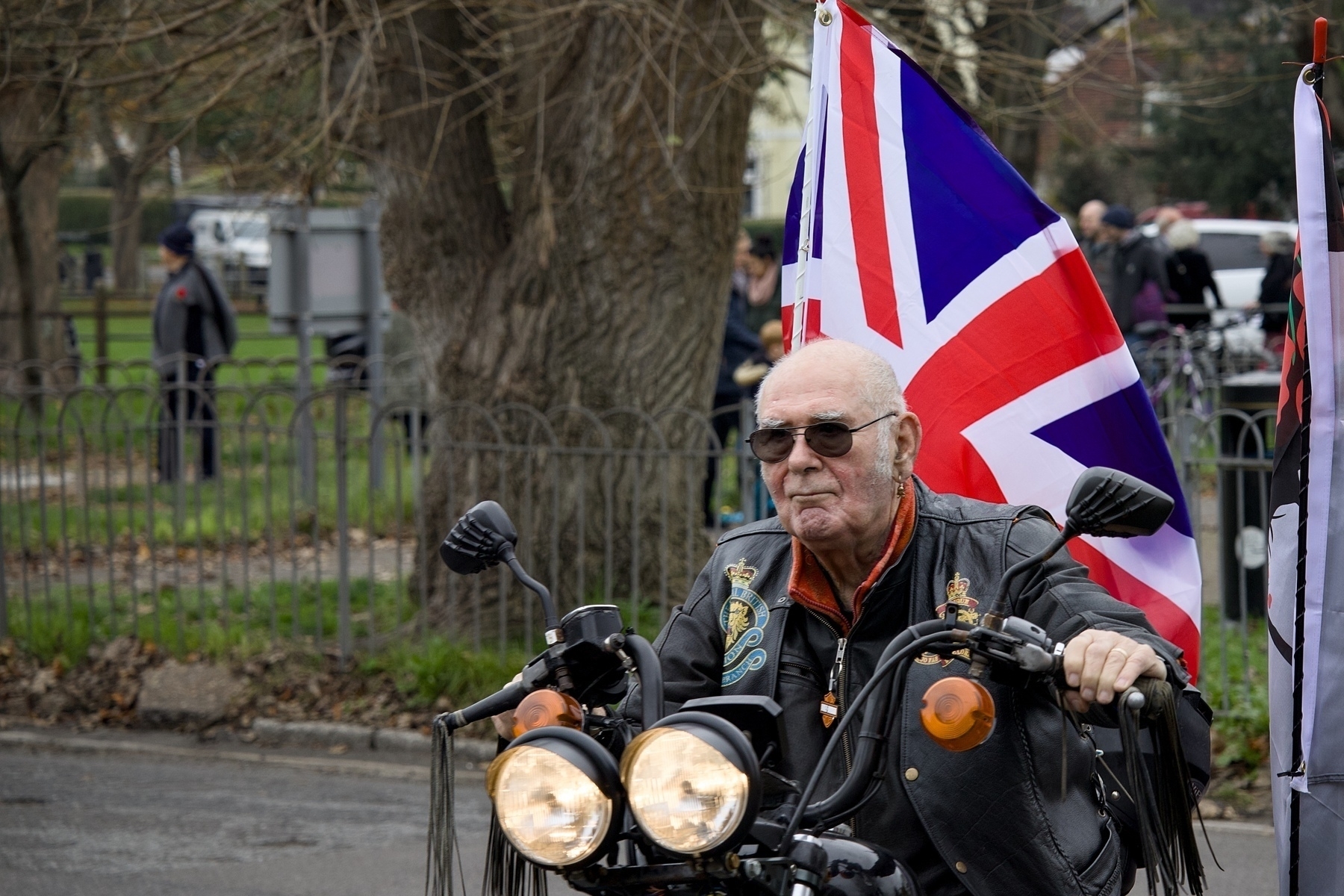 An older man is riding a motorcycle adorned with a British flag draped behind him.