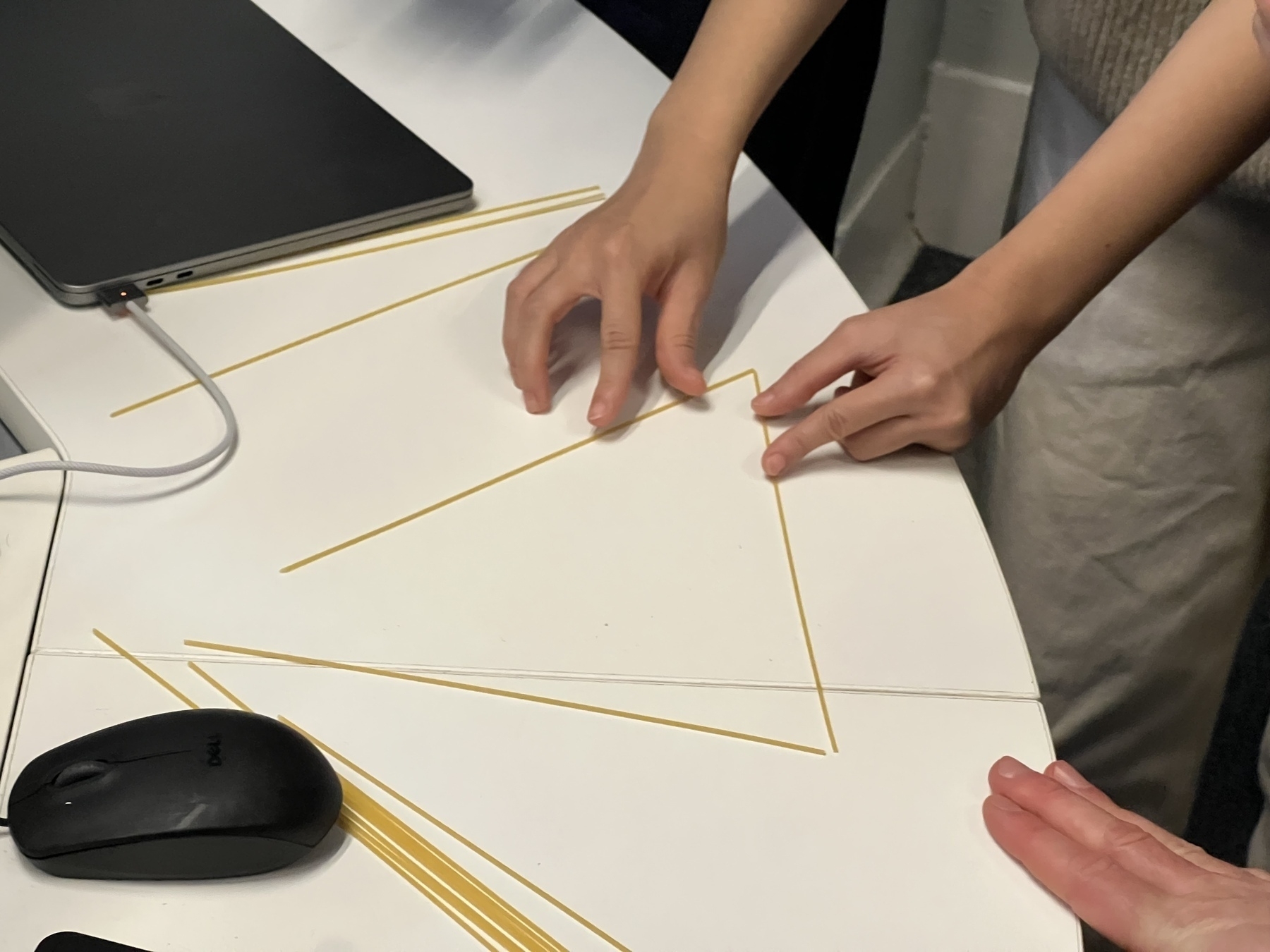 Two people are arranging uncooked spaghetti sticks into a triangular shape on a table.