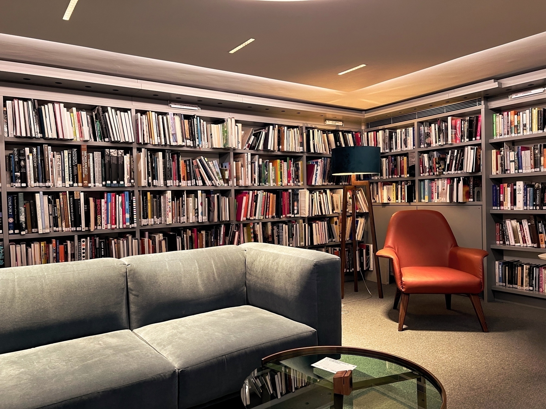 A cozy library corner features a wall of bookshelves, a gray sofa, a red armchair, and a glass coffee table.