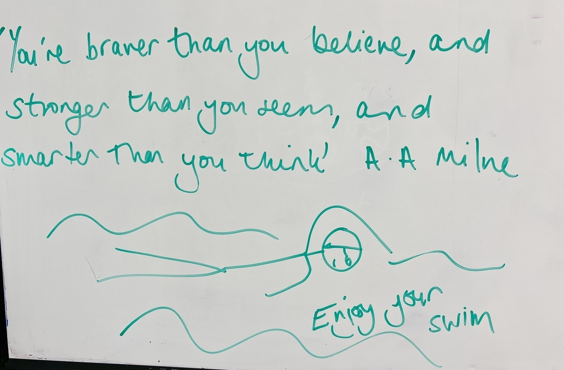 A motivational quote by A.A. Milne is written on a whiteboard, accompanied by a simple drawing of a person swimming. It reads; “You're braver than you believe, and stronger than you seem, and smarter then you think.”