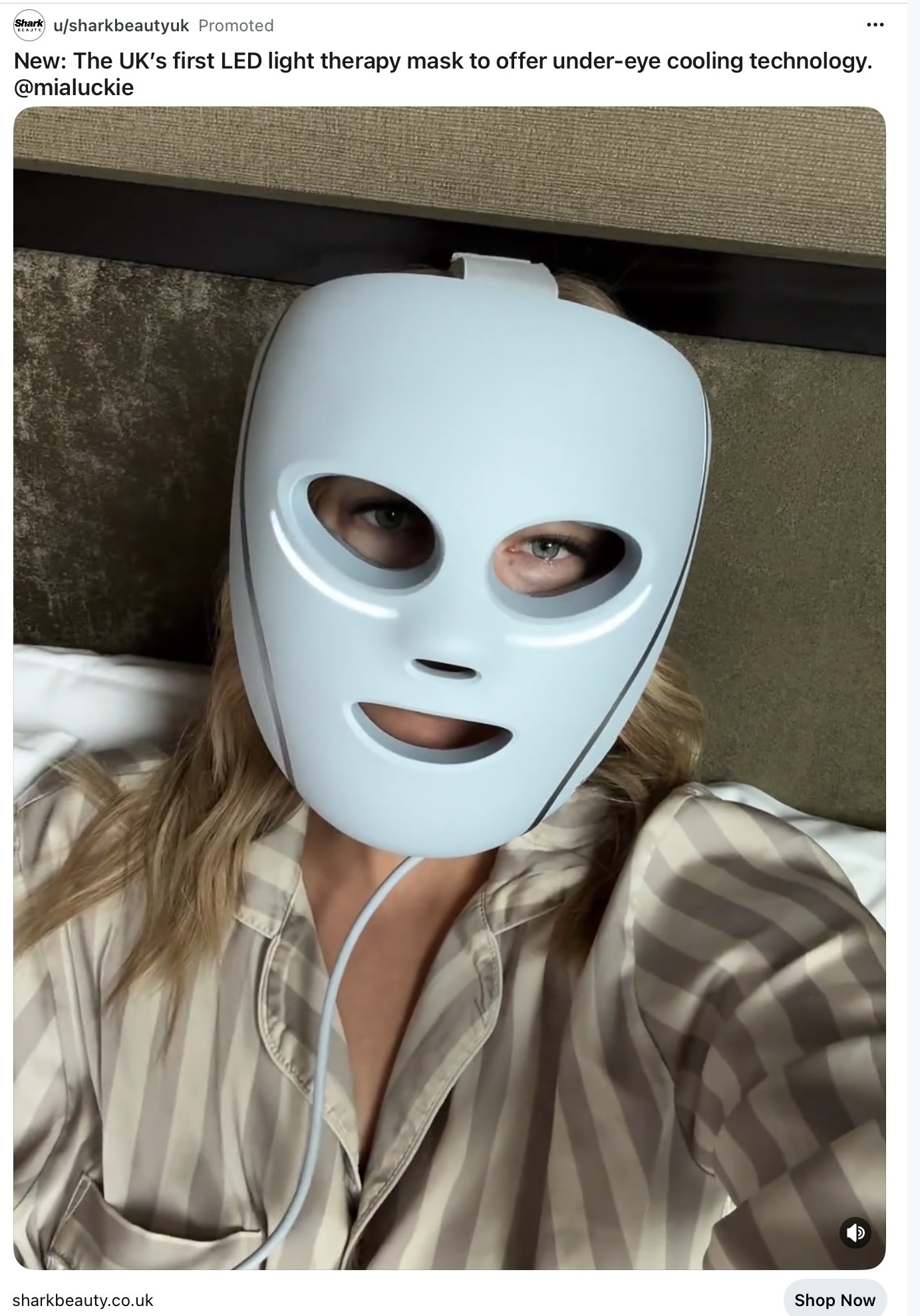 A person is wearing an LED light therapy mask with visible eye and mouth openings while lying on a bed.