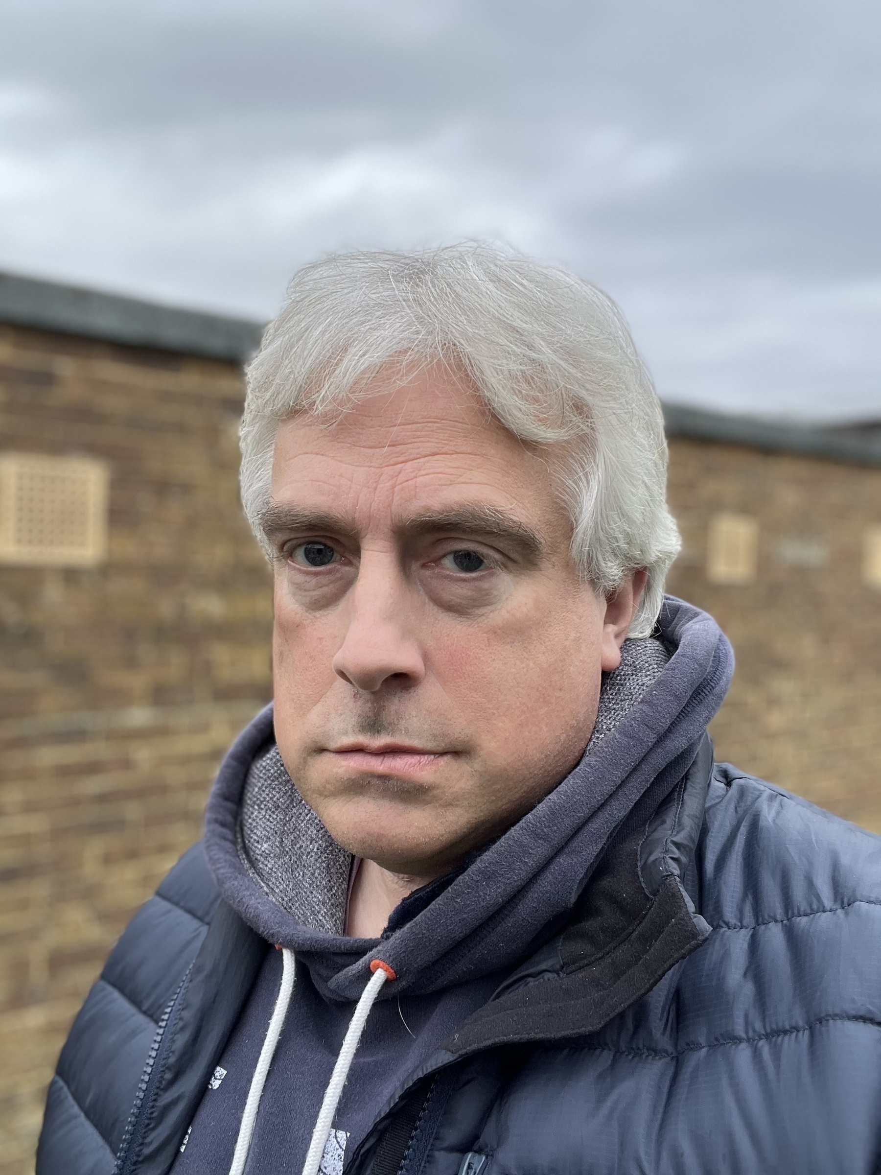 Adam Tinworth with long, unkempt hair is wearing a hoodie and jacket, standing in front of a brick wall with a cloudy sky above.