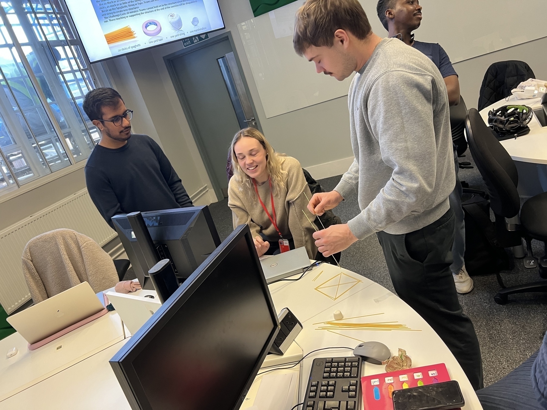 A group of people is gathered around a table working on a small structure made from sticks and tape.