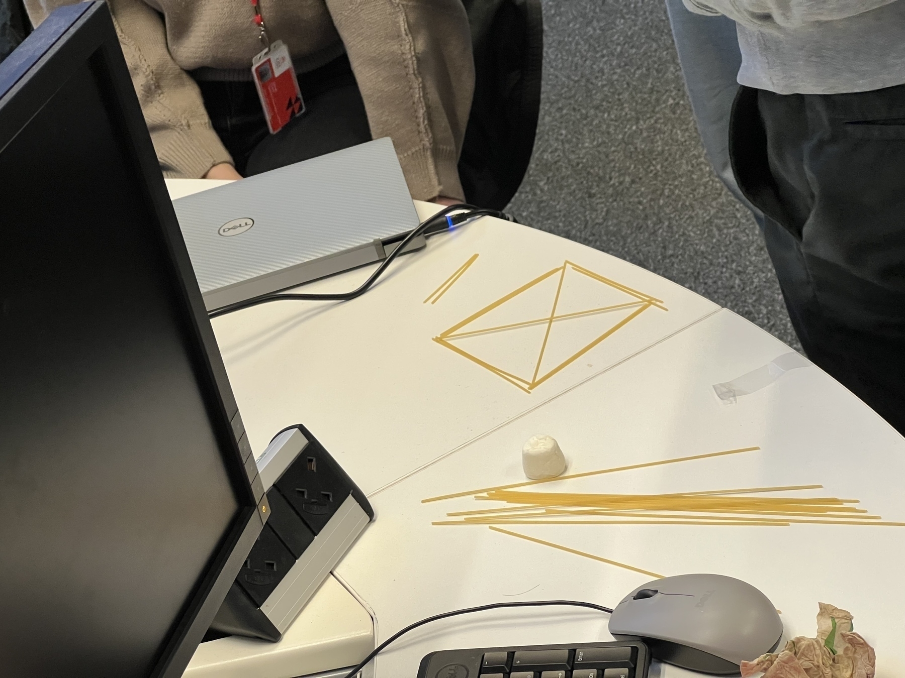 A group of people is working on a desk with a collection of uncooked spaghetti, a marshmallow, and some electronic devices and accessories nearby.