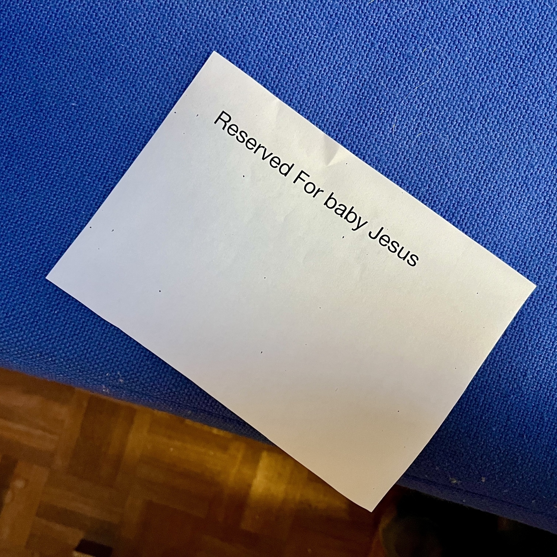 A small white paper with the text "Reserved For baby Jesus" rests on a blue fabric church pew. 